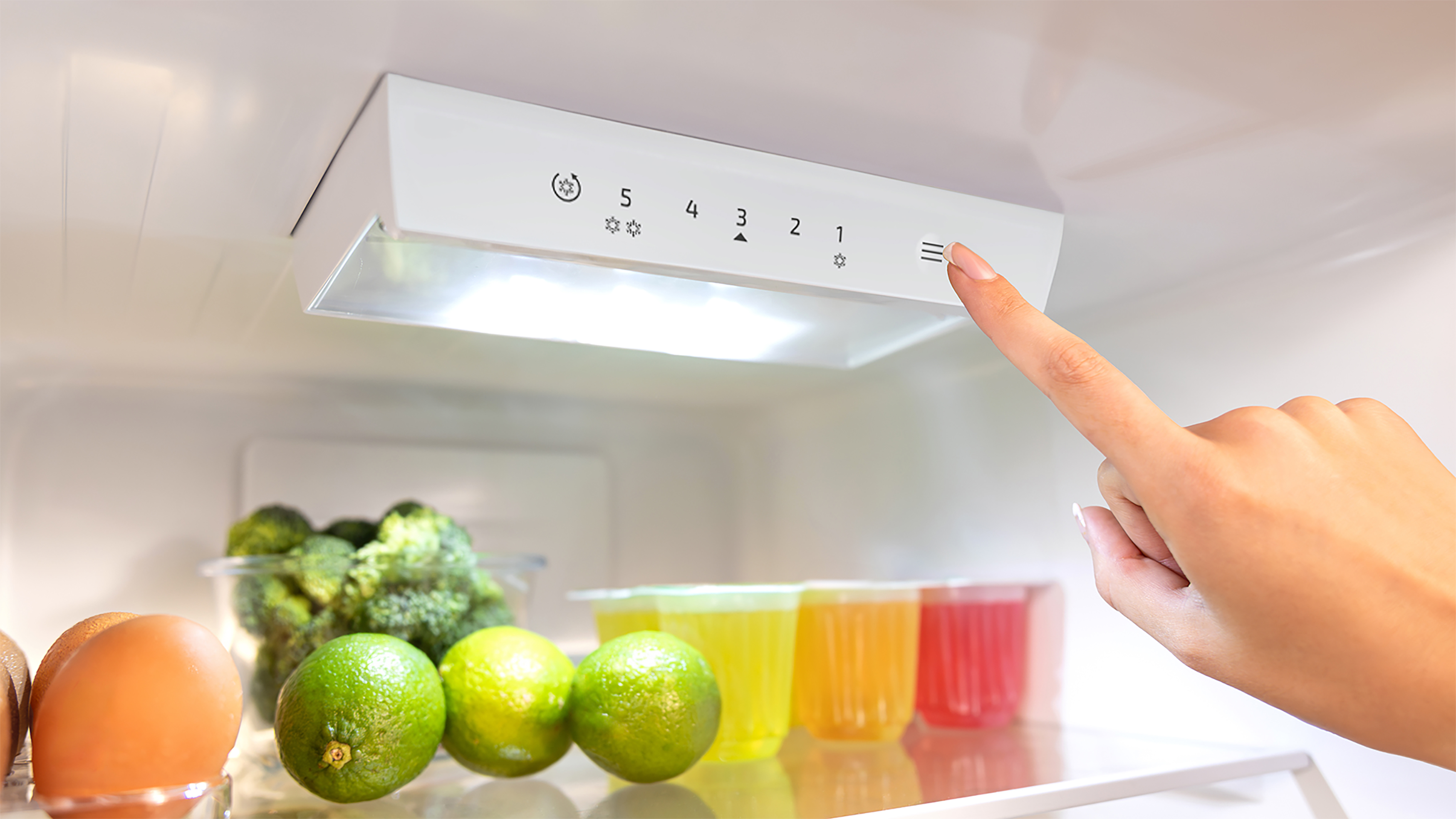 Mano tocando controles del refrigerador. Dentro, brócoli, limas y bebidas visibles.