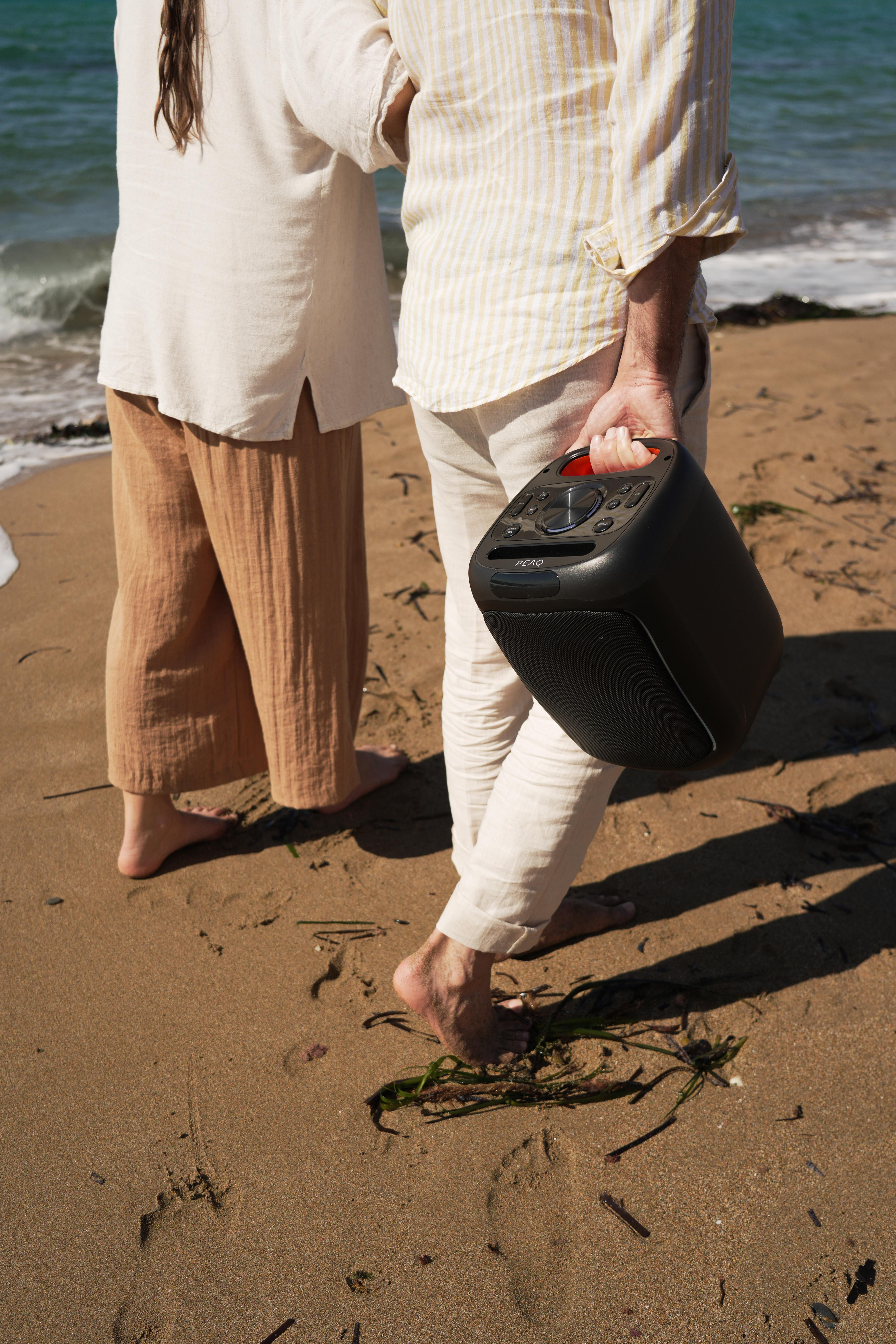 Zwei Personen am Strand, eine hält einen schwarzen Lautsprecher.