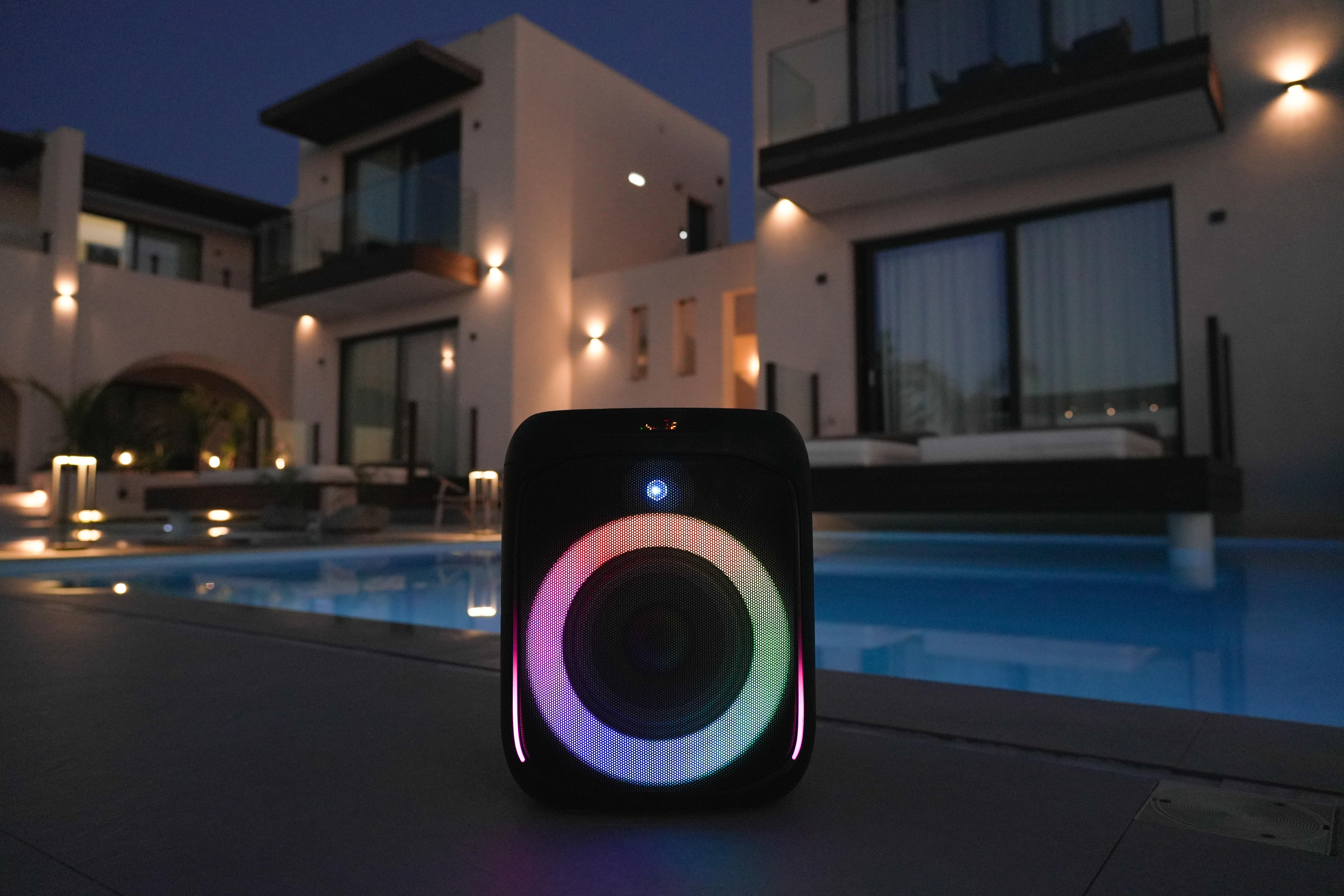 Ein schwarzer Outdoor-Lautsprecher in der Nähe eines Pools bei Nacht. Er hat einen farbenfrohen Lichtring.