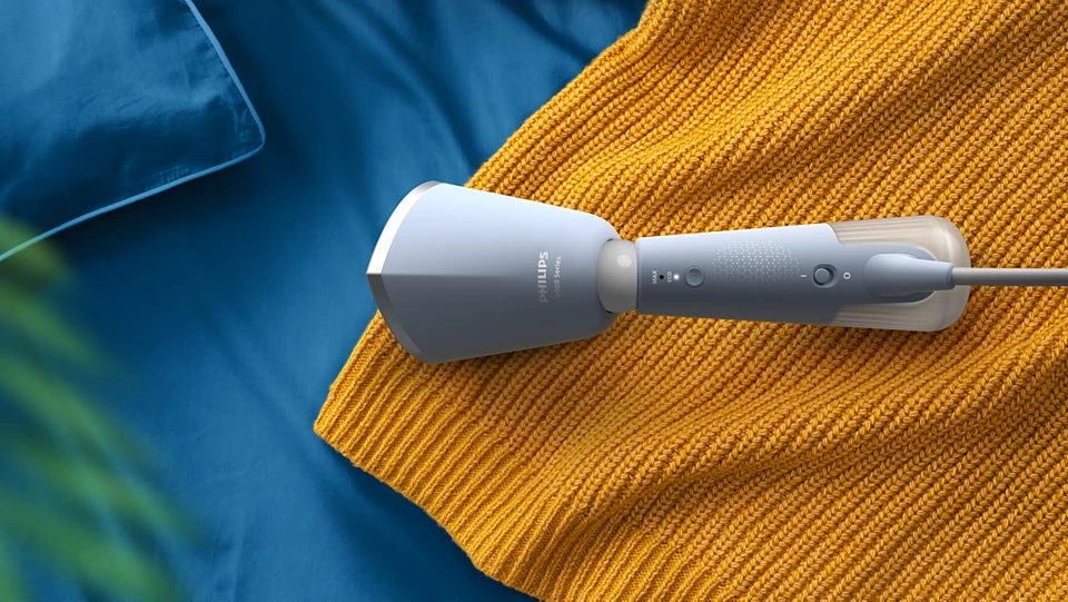 Un défroisseur Philips gris sur un pull orange, avec un fond d'oreiller bleu.