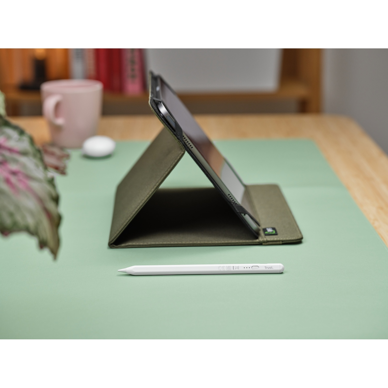 Una custodia per tablet color oliva con una penna su una superficie verde, con una tazza rosa e libri sullo sfondo.