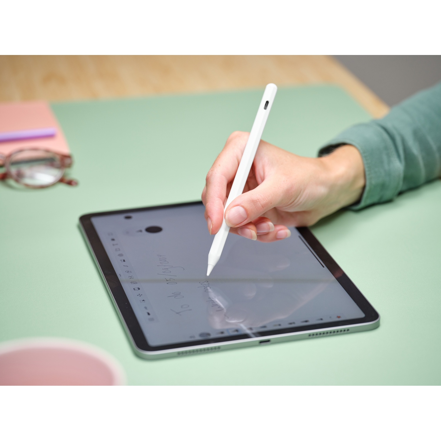 Una mano scrive su un tablet con una penna bianca. Scrivania verde, blocco note rosa.