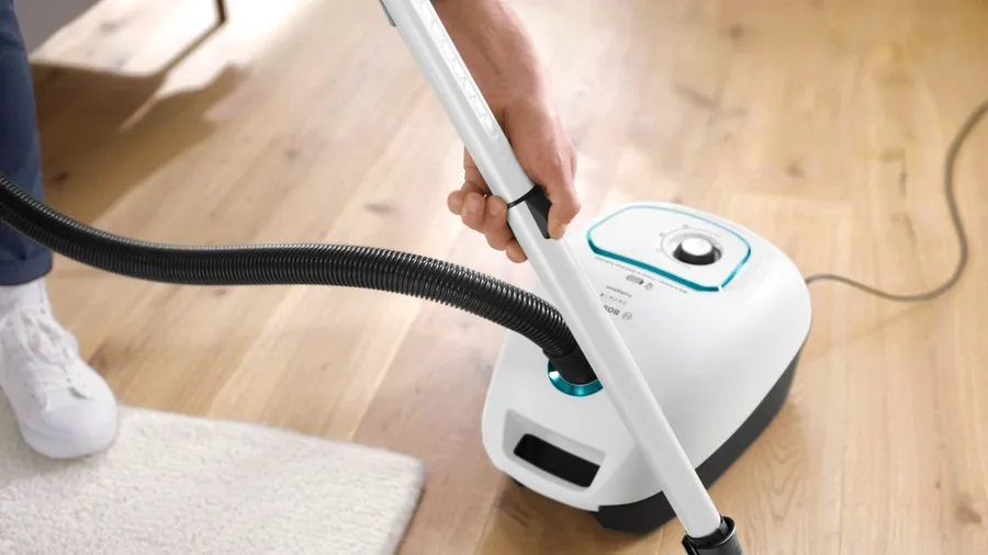 Osoba używa białego i turkusowego odkurzacza na drewnianej podłodze. Biały dywan jest na pierwszym planie.