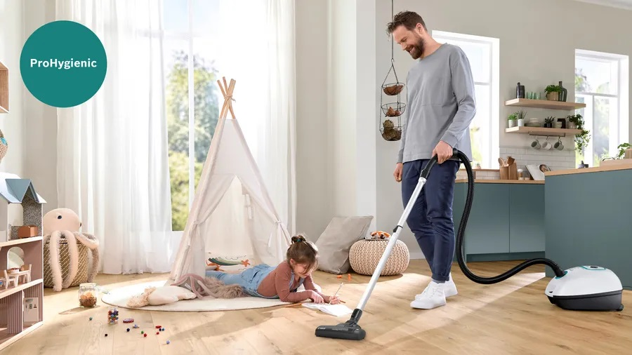 Mężczyzna odkurza podłogę, podczas gdy dziecko rysuje w namiocie. Widoczny jest odkurzacz.