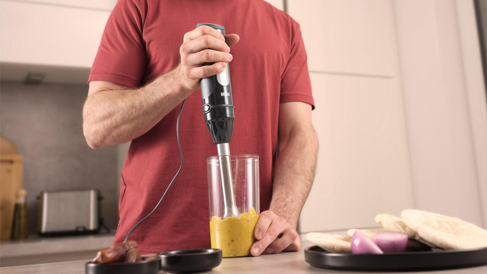 Person verwendet Stabmixer in einem Glas mit gelben Zutaten, Kochszene.