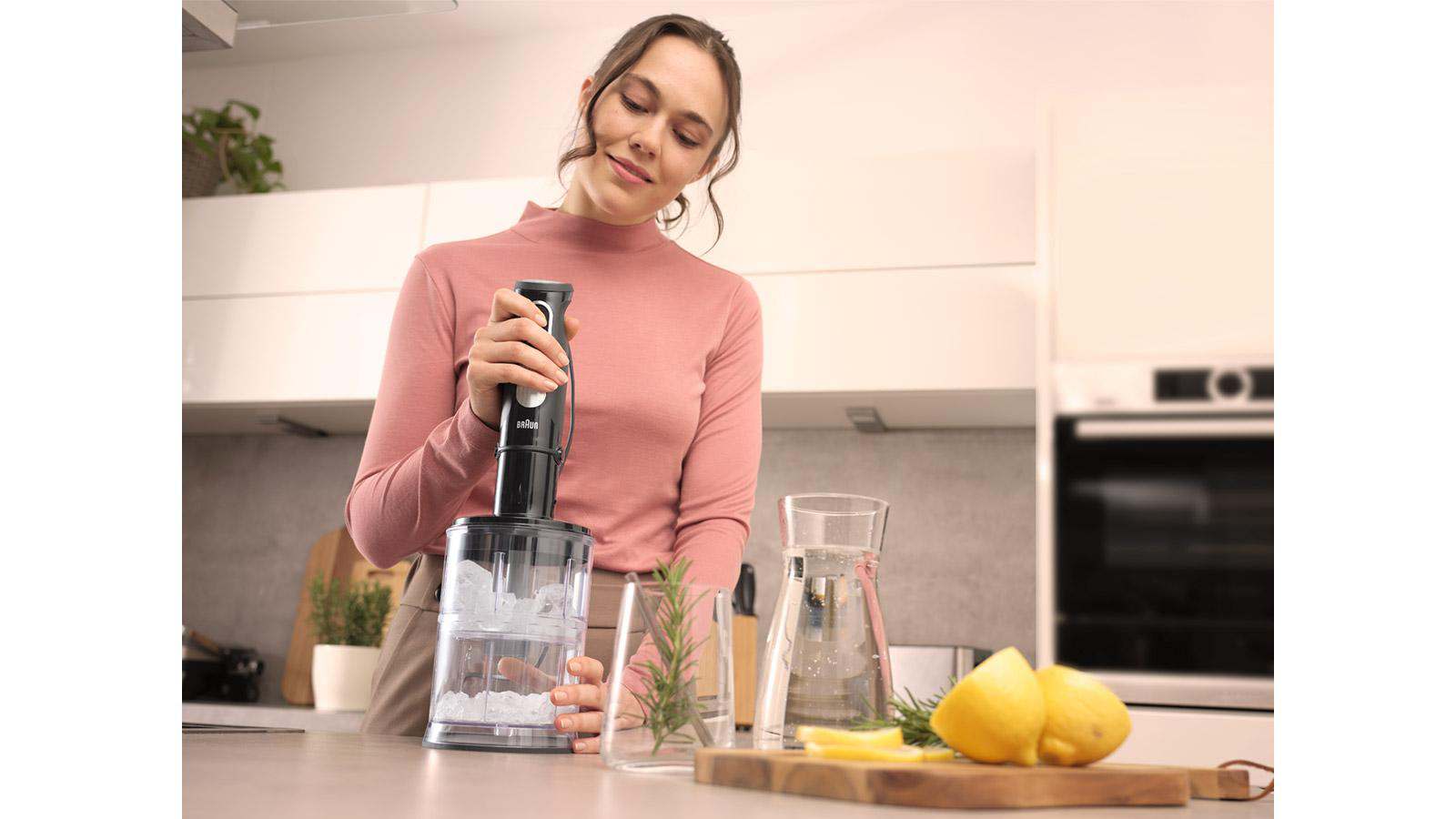 Eine Frau verwendet einen Stabmixer, um Zutaten in einem Behälter zu mischen, mit Obst und Kräutern.