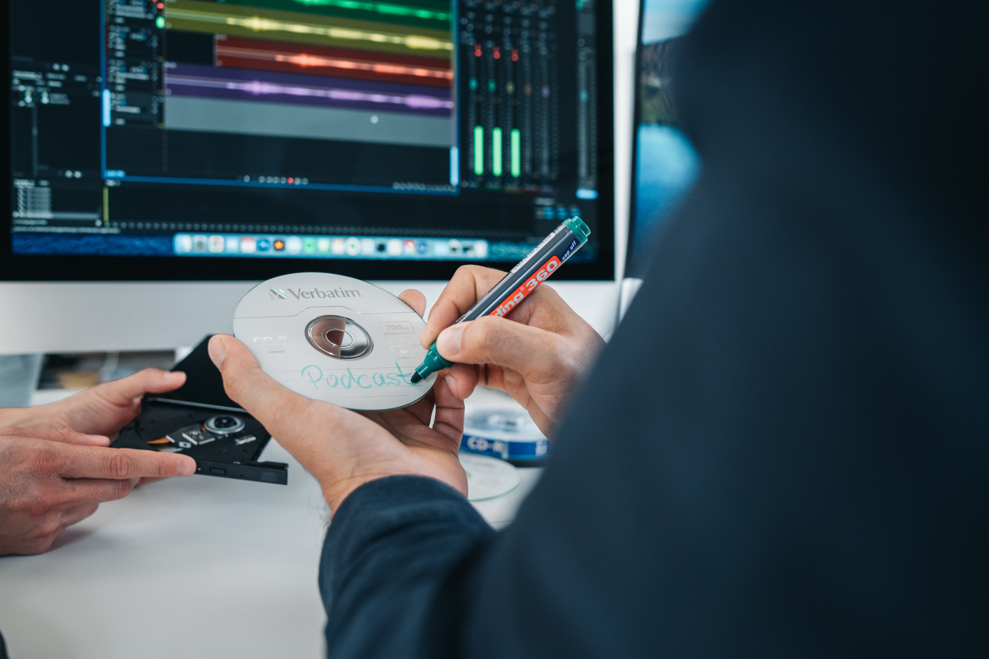 Dwie ręce piszące 'Podcast' na płycie CD. Ekran komputera i inne płyty CD w tle.