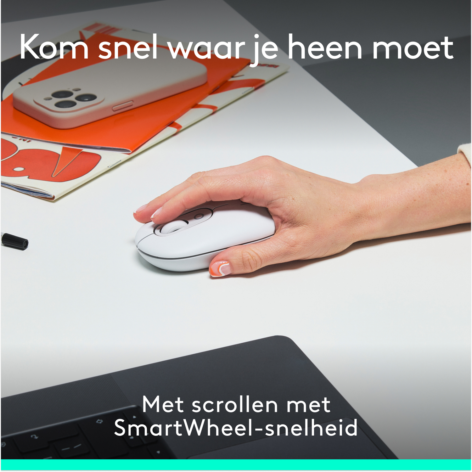 Hand gebruikt witte muis op witte tafel, laptop op de achtergrond. Notitieboek en telefoon liggen op de tafel.