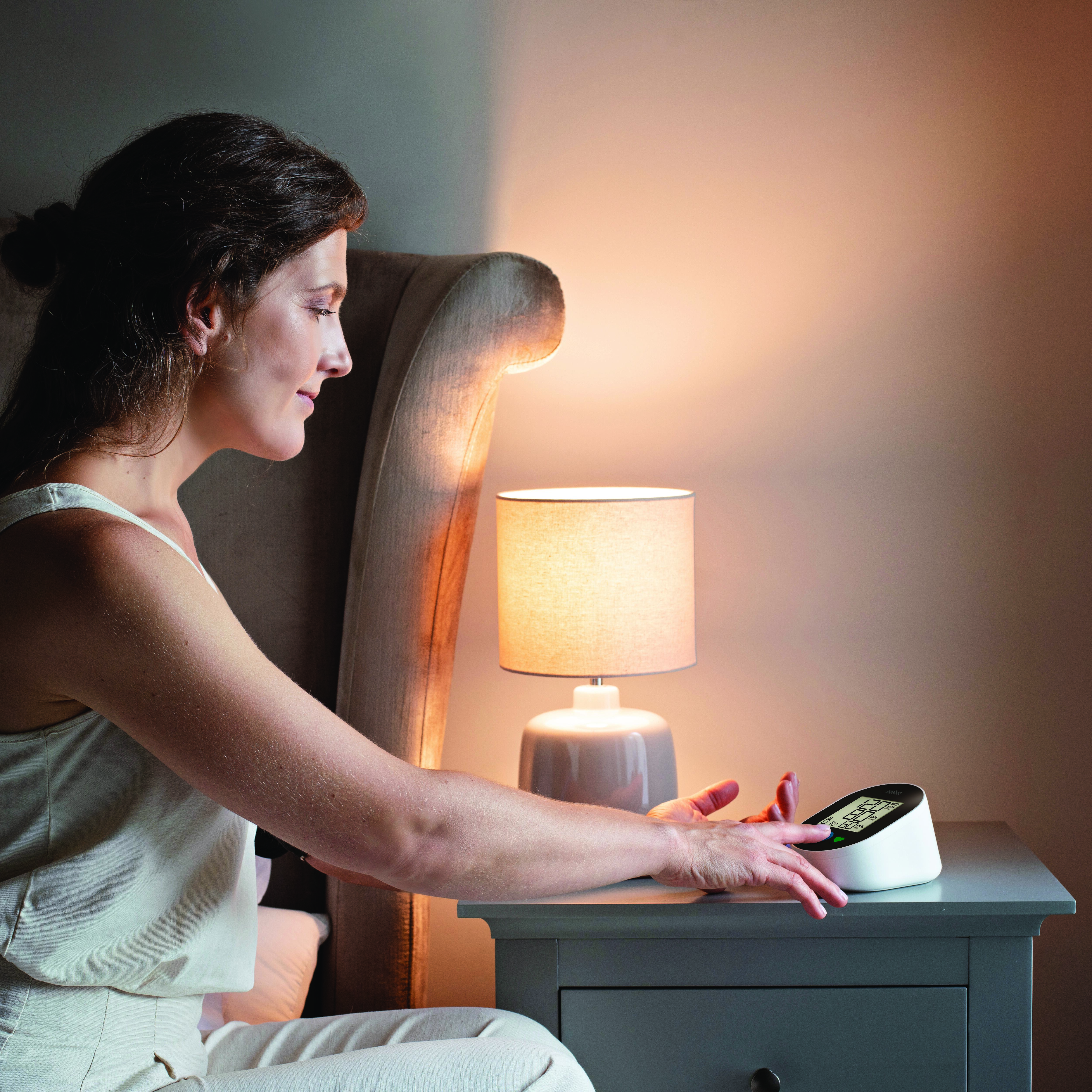 Eine Frau benutzt ein Blutdruckmessgerät auf einem Nachttisch neben einer Lampe.