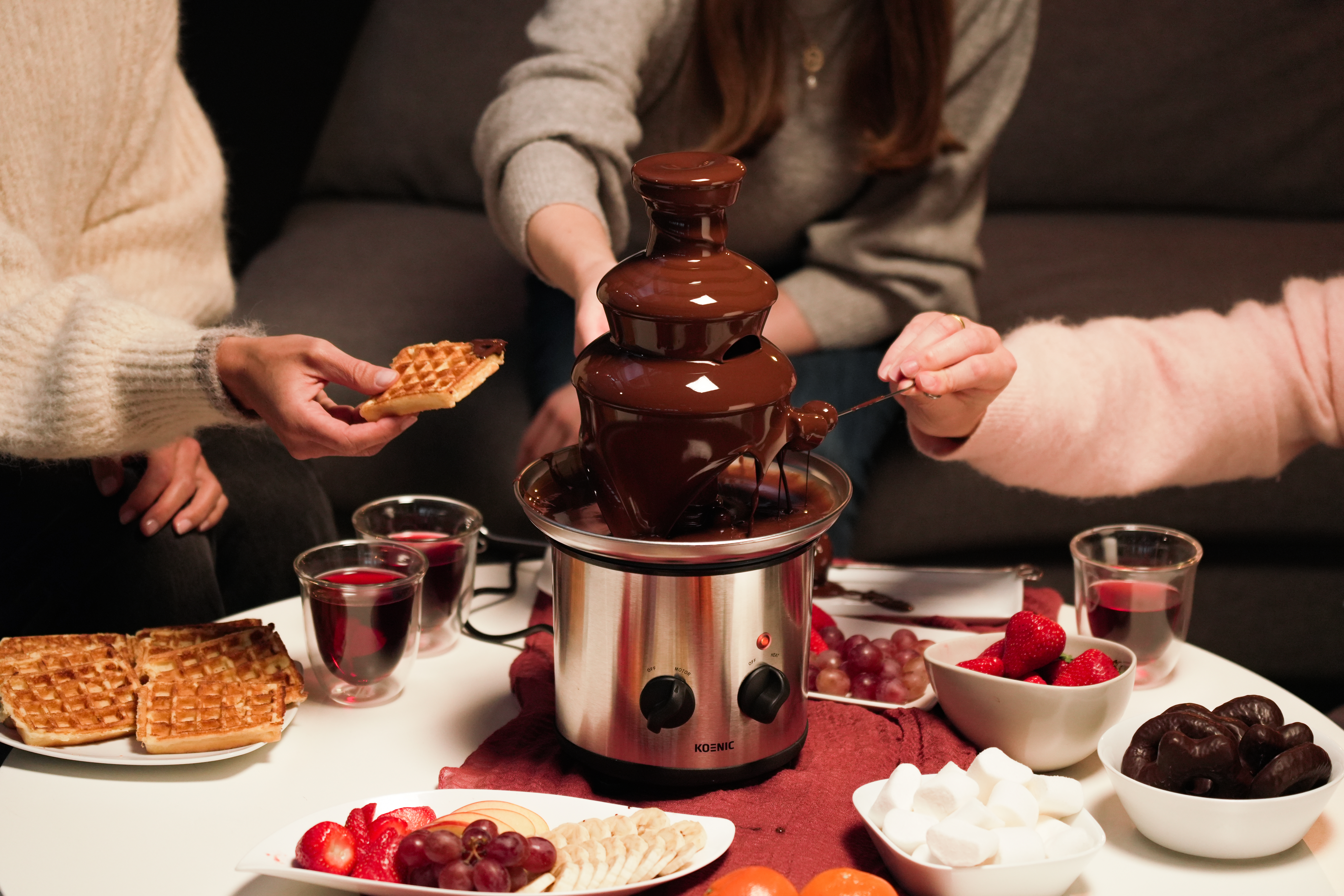 Personen um einen Schokoladenbrunnen mit Waffeln, Obst und Süßigkeiten.