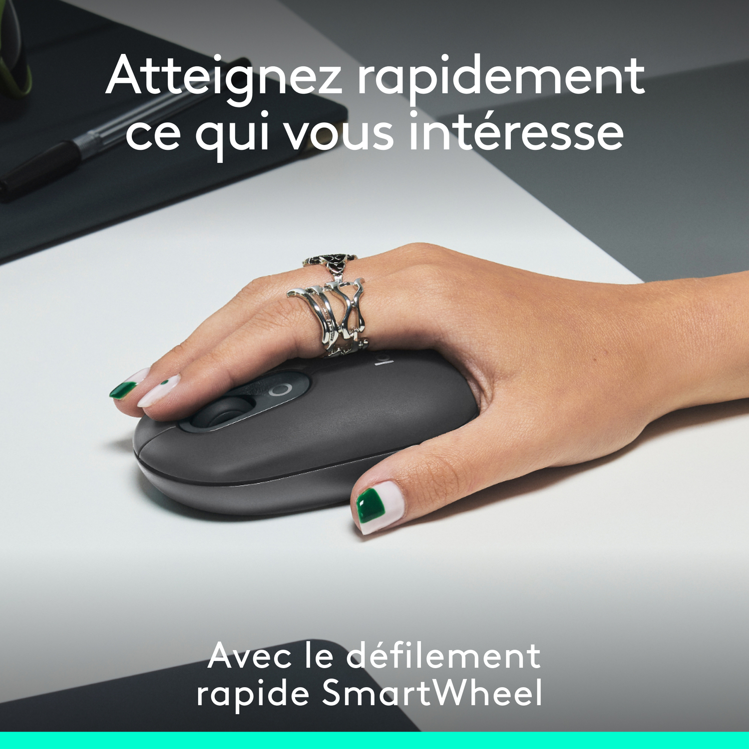 Une main avec des bagues utilisant une souris gris foncé sur un bureau blanc.