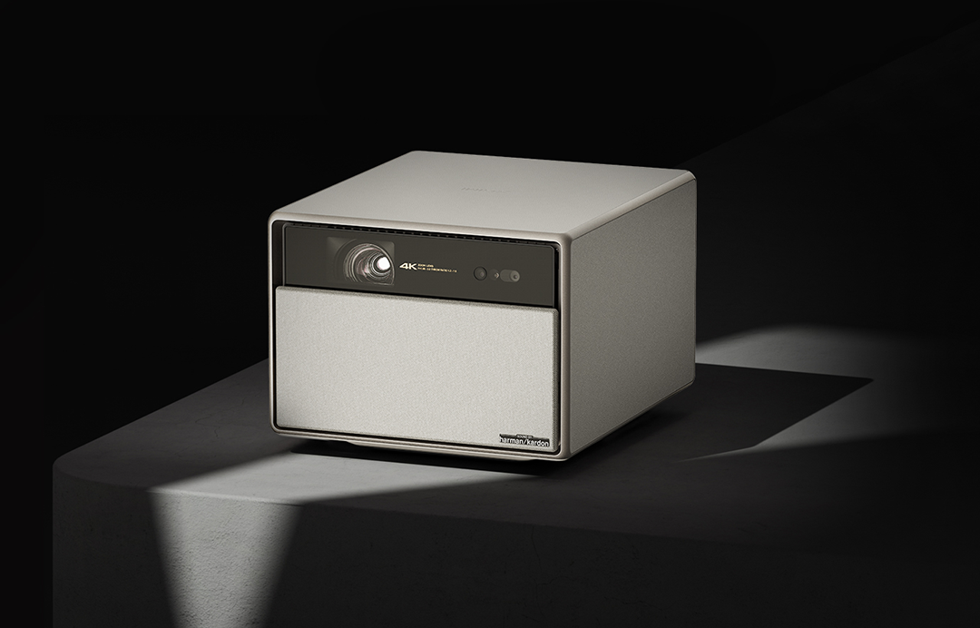 Fehér és szürke projektor fehér platformon. Fekete háttér. 'Harman Kardon' logó.