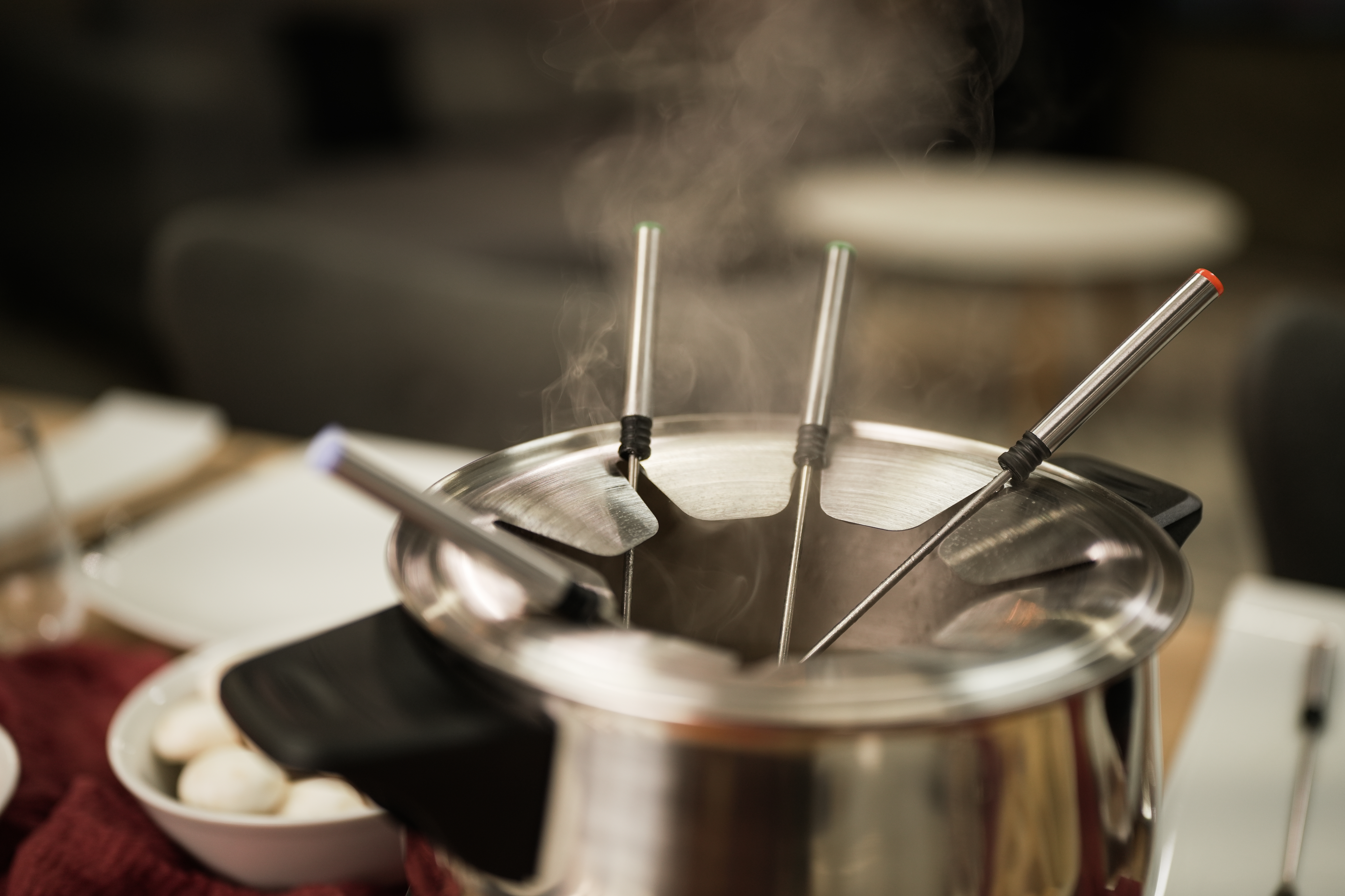 Ein Fonduetopf mit Spießen, Dampf steigt auf, auf einem Tisch. Weicher Fokus, unscharfer Hintergrund.
