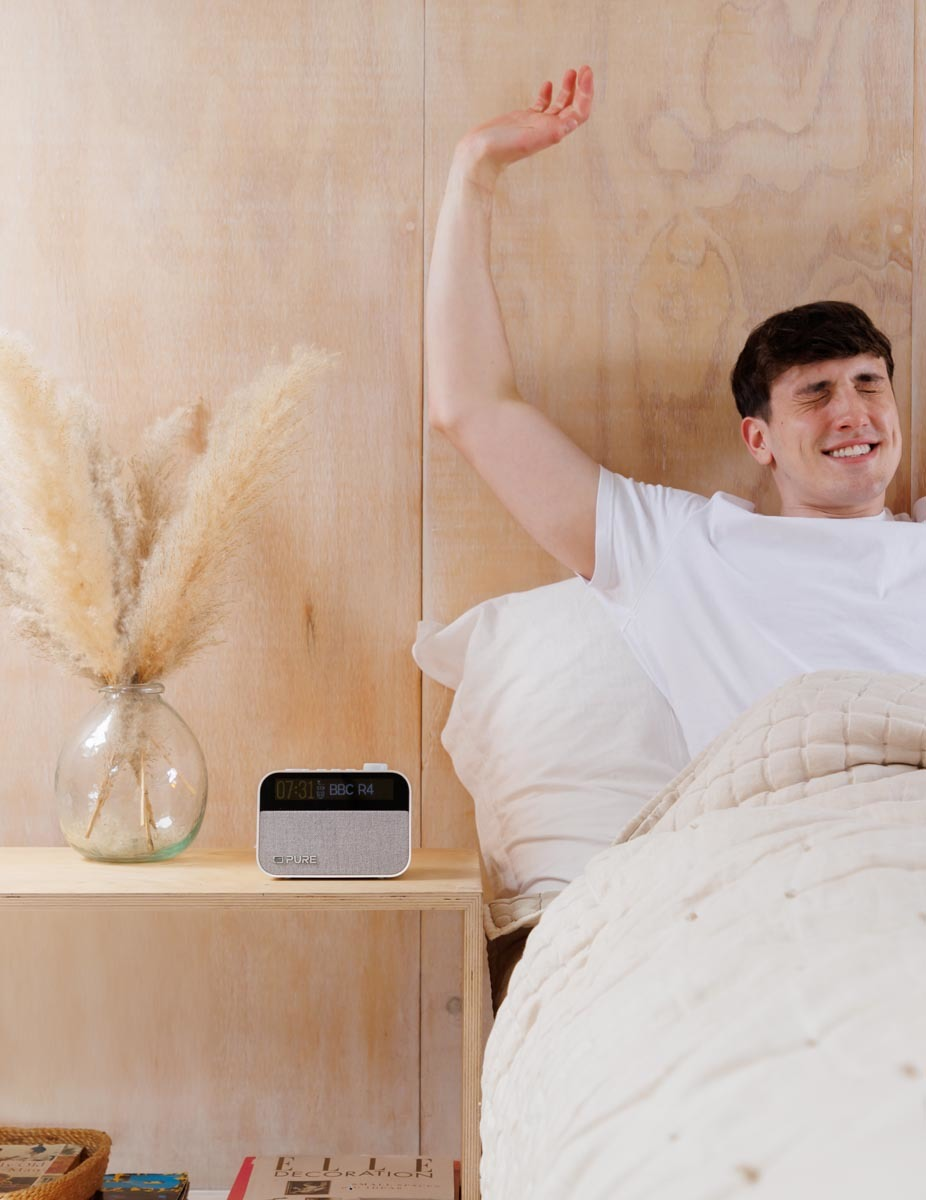 Un homme au lit s'étire, avec une radio et un vase sur la table de chevet. Tons beiges dominent la pièce.
