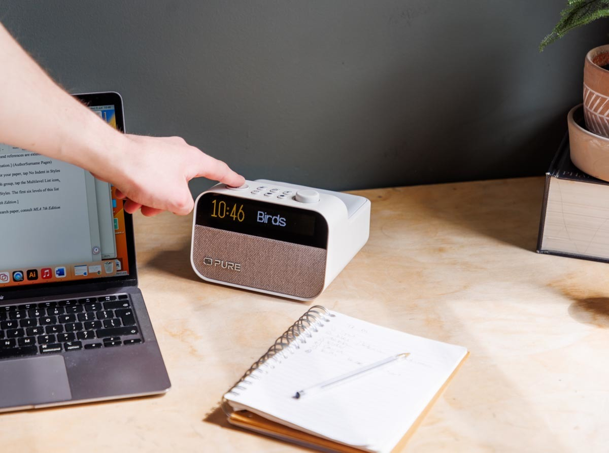 Main appuyant sur un bouton d'un radio-réveil blanc. Ordinateur portable et bloc-notes sur le bureau.