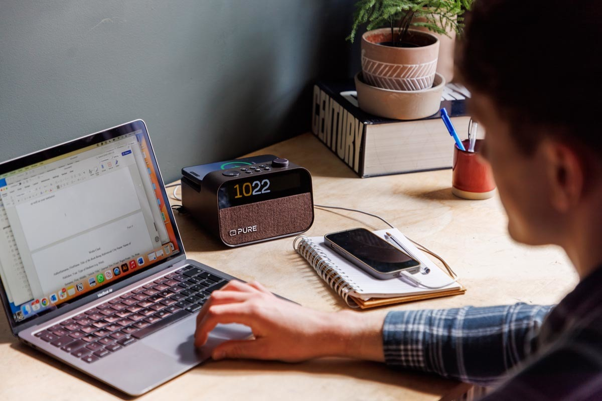 Une personne travaille sur un ordinateur portable. Une radio et un téléphone sont à côté. La scène est sur un bureau en bois.