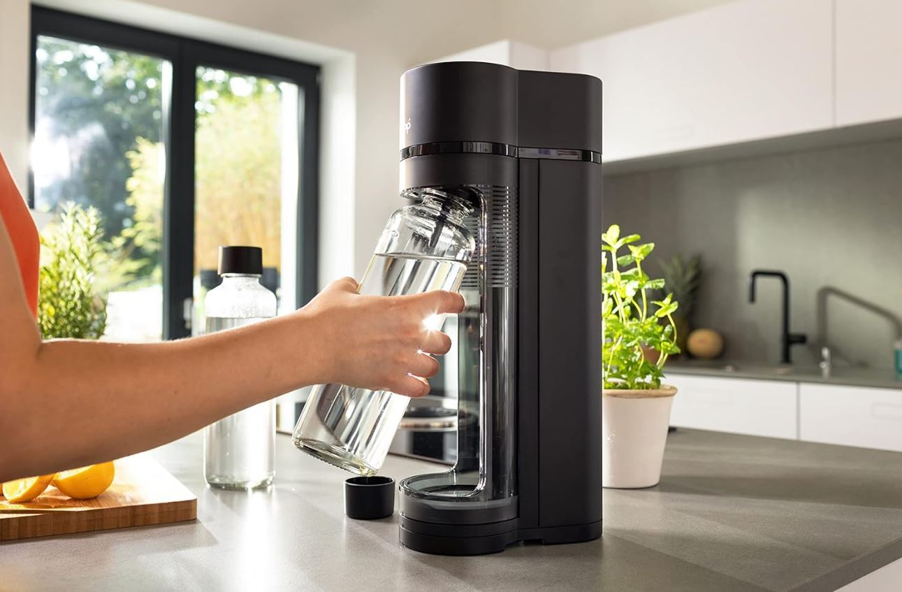 Person gießt Wasser aus einer Glasflasche in einen Wassersprudler.
