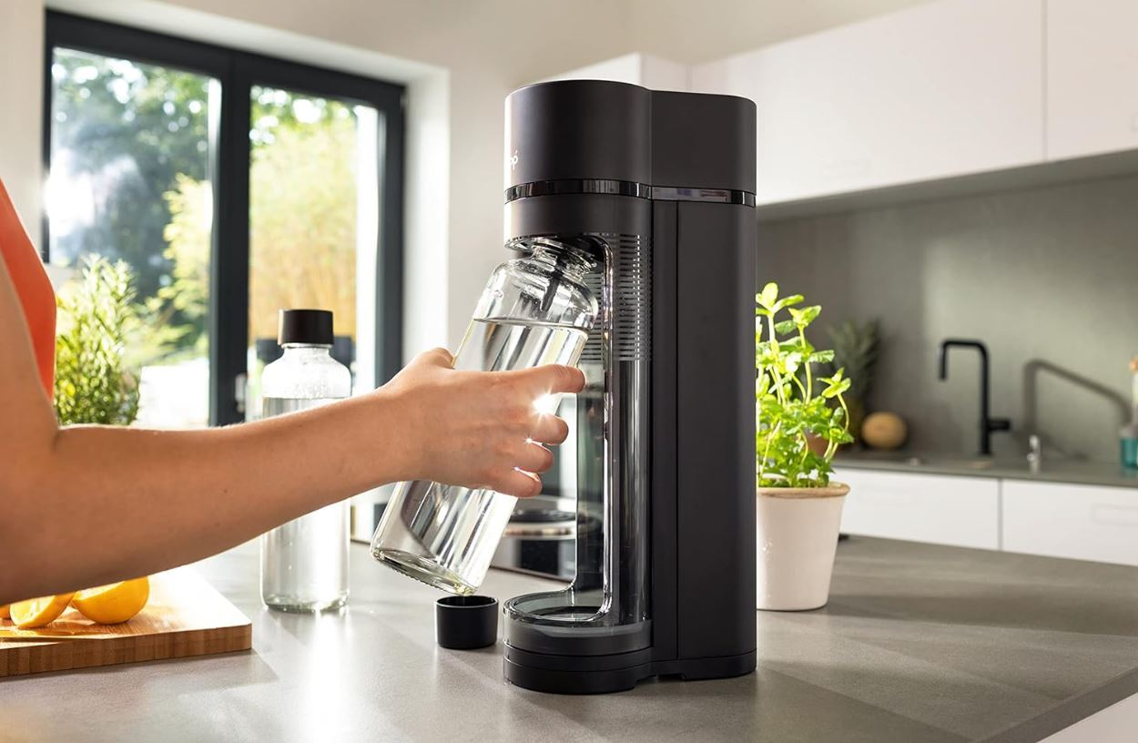 Eine Person gießt Wasser in einen schwarzen Soda-Maker mit einer Glasflasche auf einer Theke.
