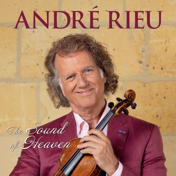André Rieu houdt een viool vast en glimlacht. Hij draagt een roze overhemd en een rode jas. Achter hem een stenen muur.
