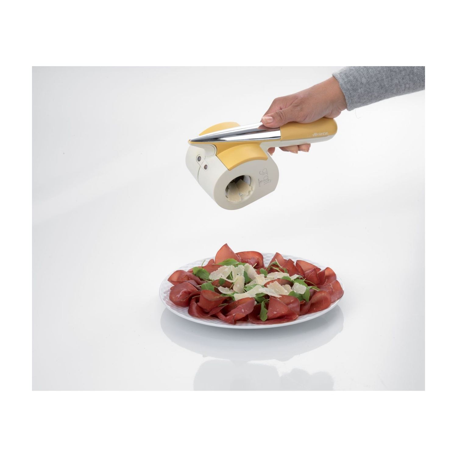 Mano che tiene una grattugia per formaggio sopra un piatto di cibo. La grattugia è gialla e bianca.