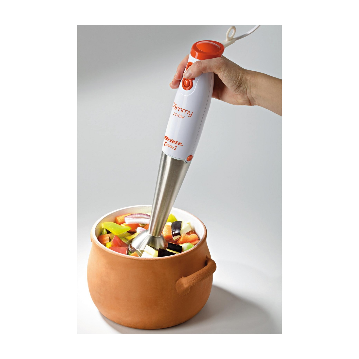 Una persona che frulla cibo in una pentola di terracotta con un frullatore a immersione. Le verdure sono visibili.