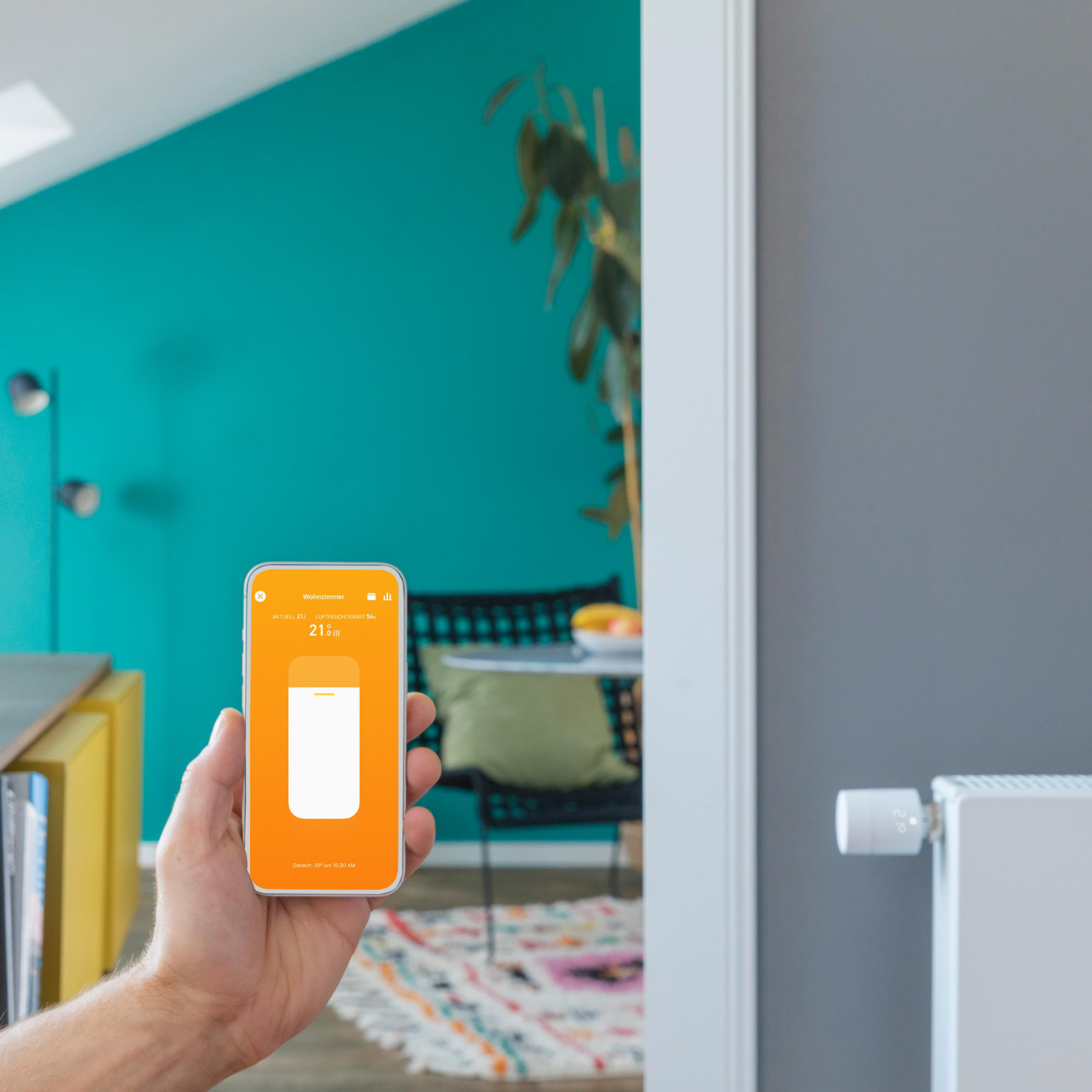 Eine Person hält ein Telefon mit Smart-Home-App und Temperatureinstellung. Ein Heizkörper im Hintergrund.