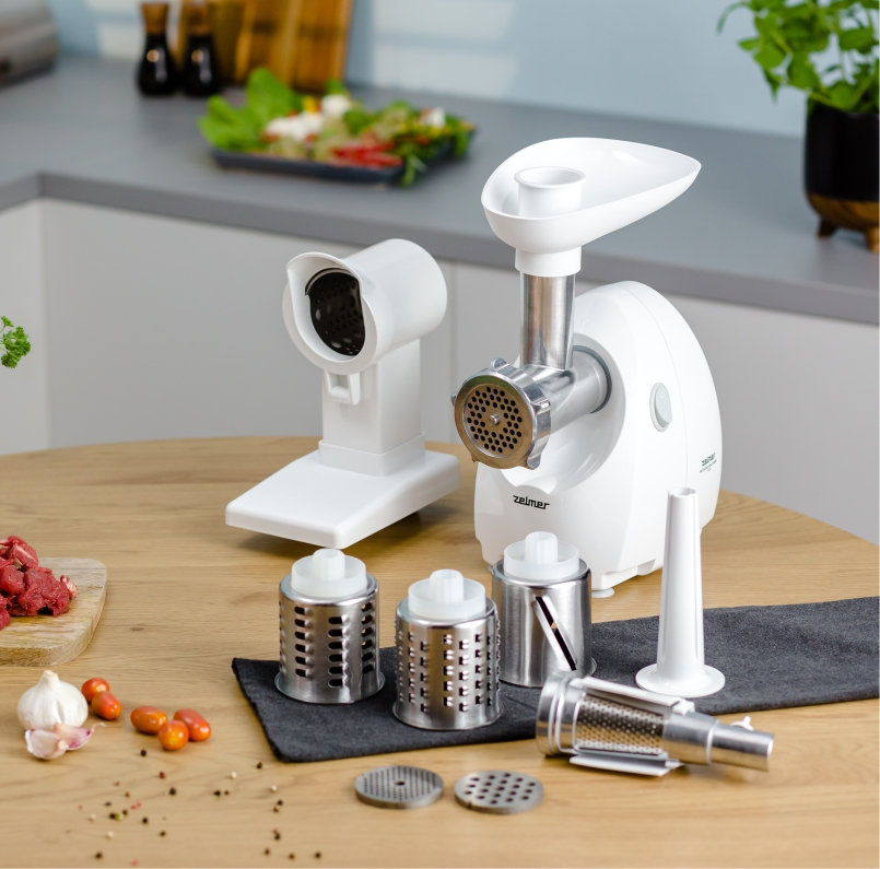 Biała maszynka do mięsa z przystawkami na drewnianym stole. Warzywa i mięso również na stole.