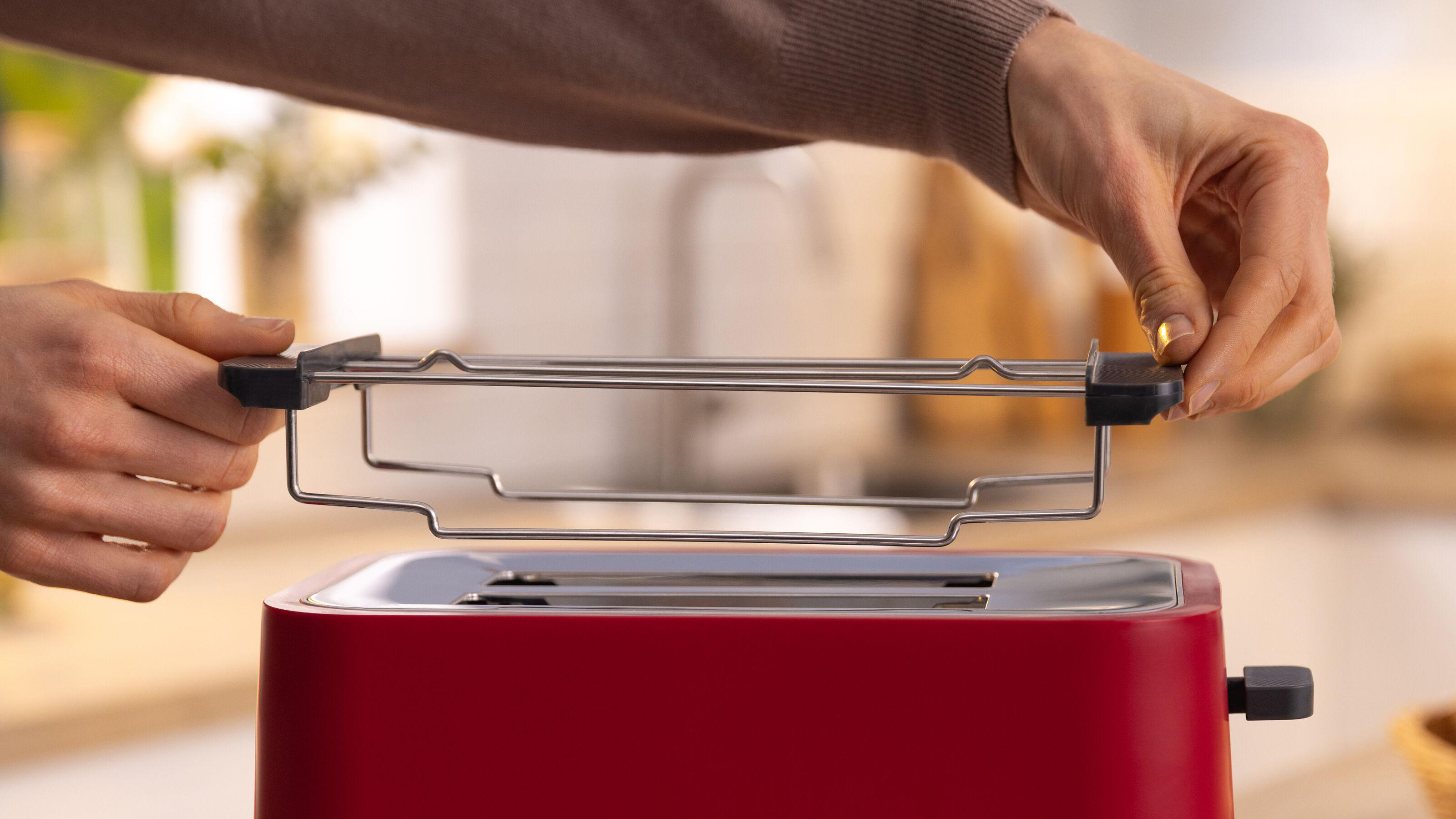 Hände, die ein Metallgestell in einen roten Toaster setzen, auf einer hellen Küchenarbeitsplatte.