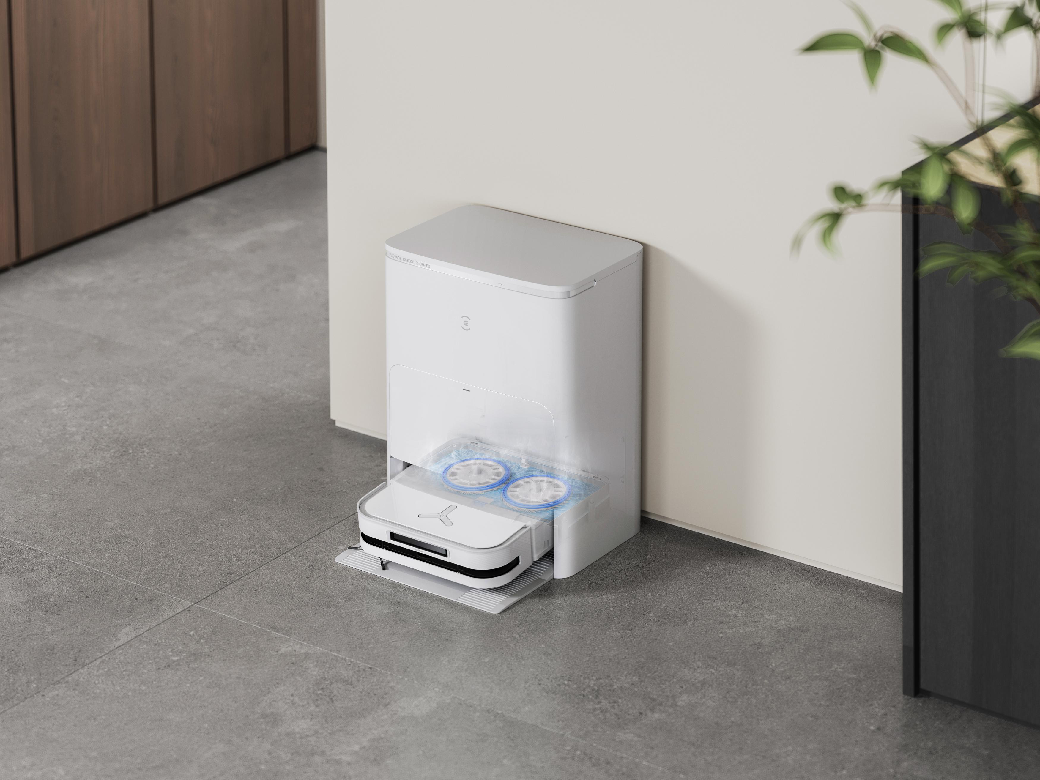 Ein weißer Roboter-Staubsauger mit einer Ladestation. Er steht auf einem grauen Fliesenboden.