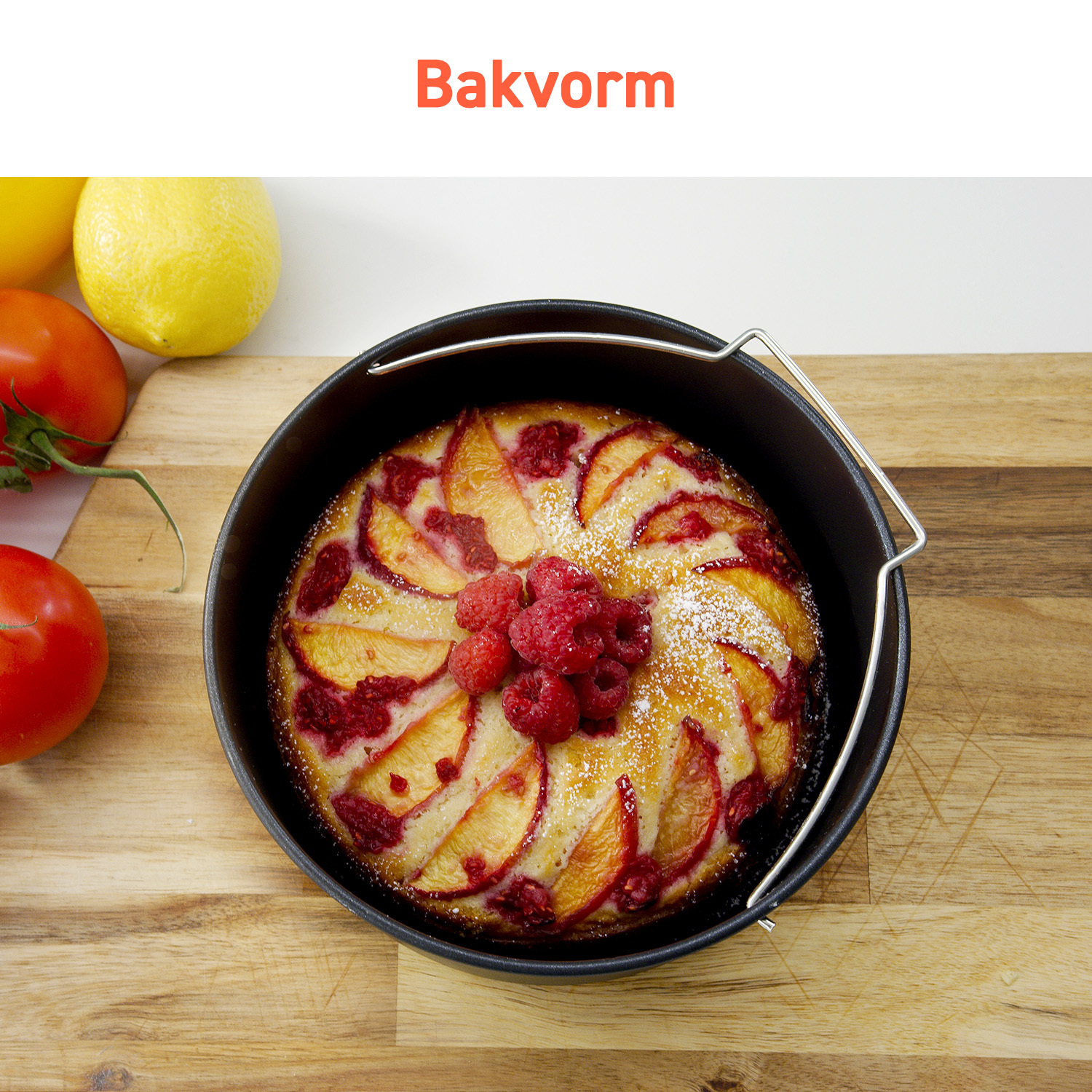 A cake in a black baking form, topped with peaches and raspberries, beside tomatoes and lemons on a wooden surface.