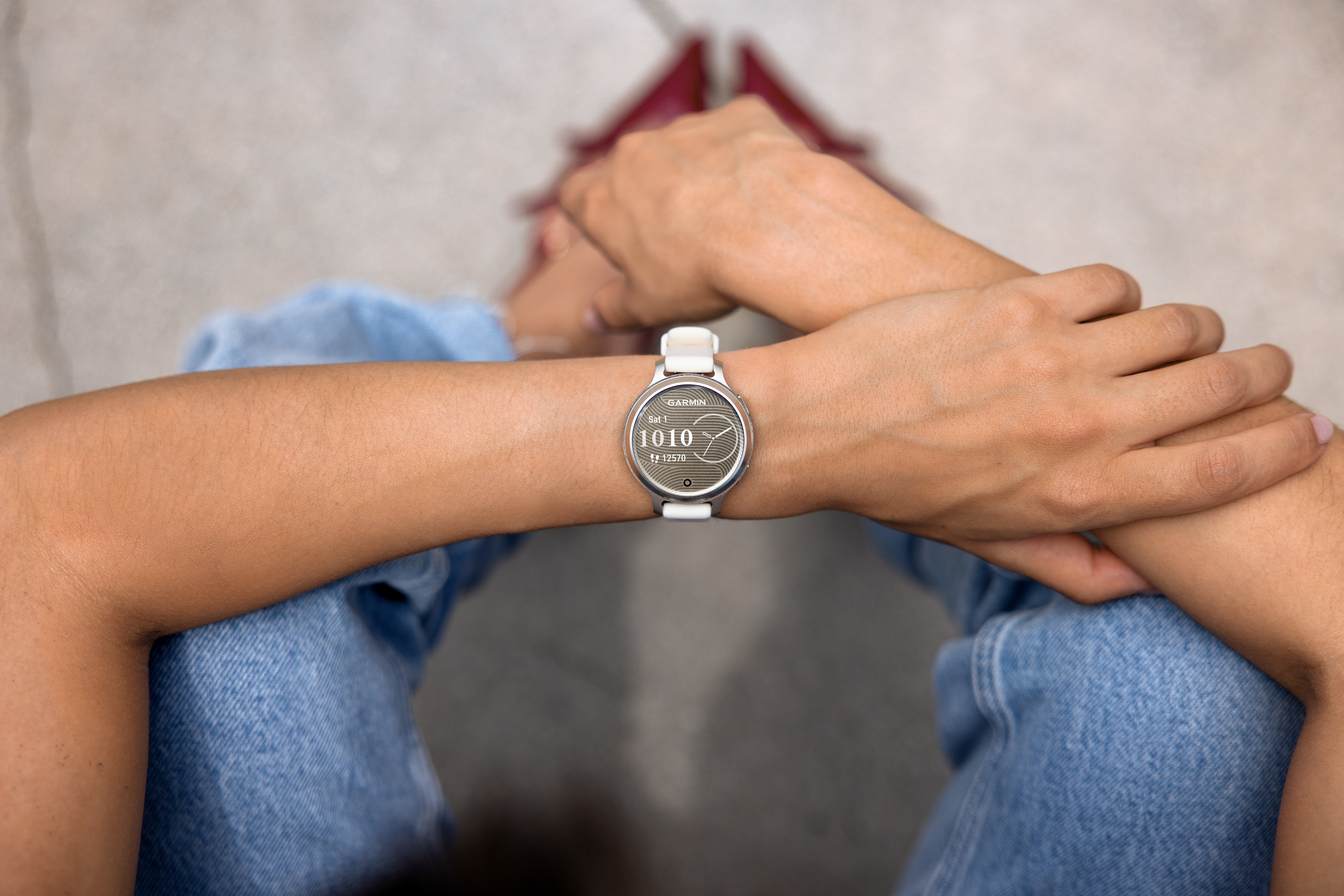 Une personne porte une smartwatch Garmin. La montre affiche l'heure et les données d'activité. Un jean et la peau sont visibles.