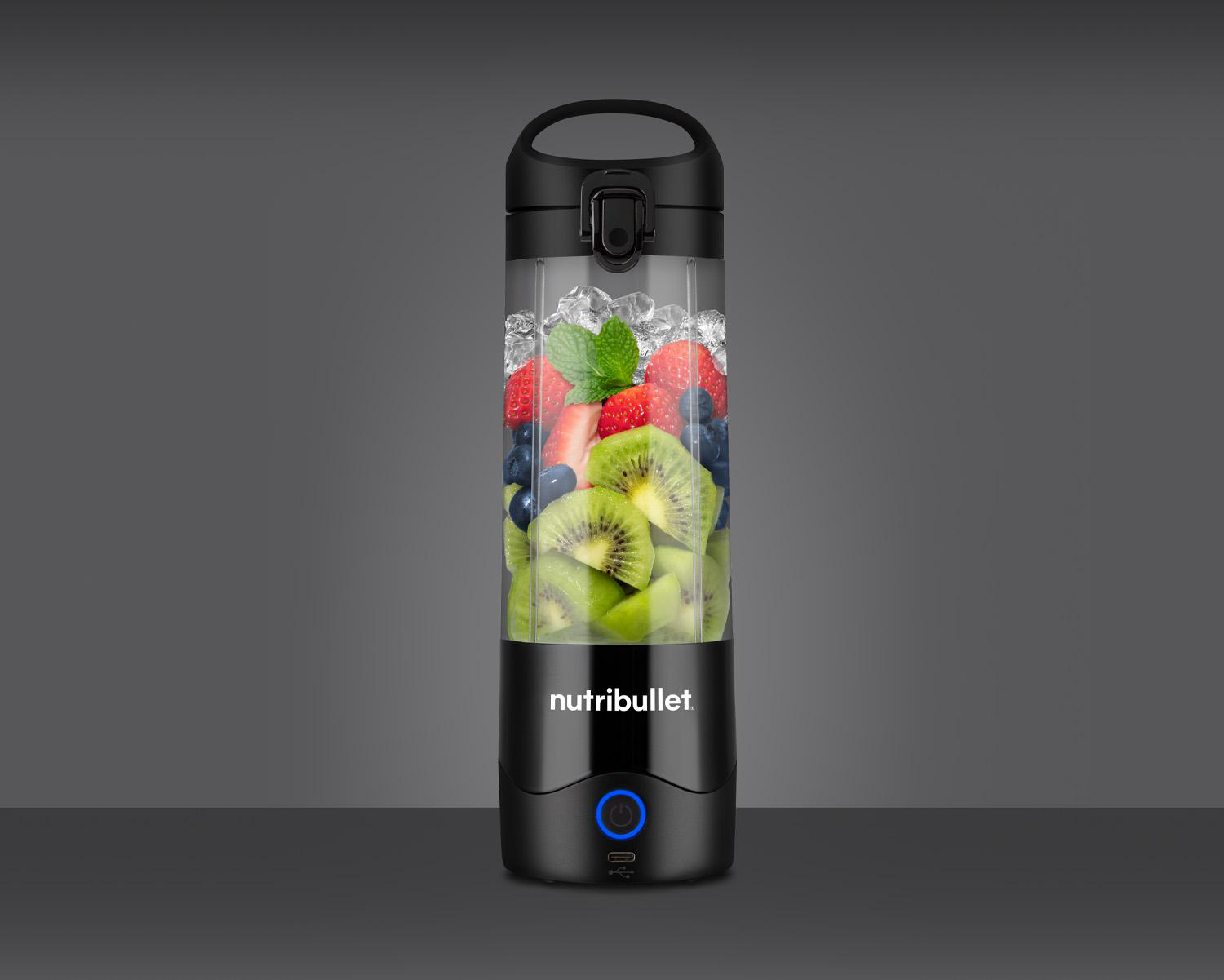 Eine Nutribullet-Blenderflasche gefüllt mit Eis, Früchten und Minze vor grauem Hintergrund.