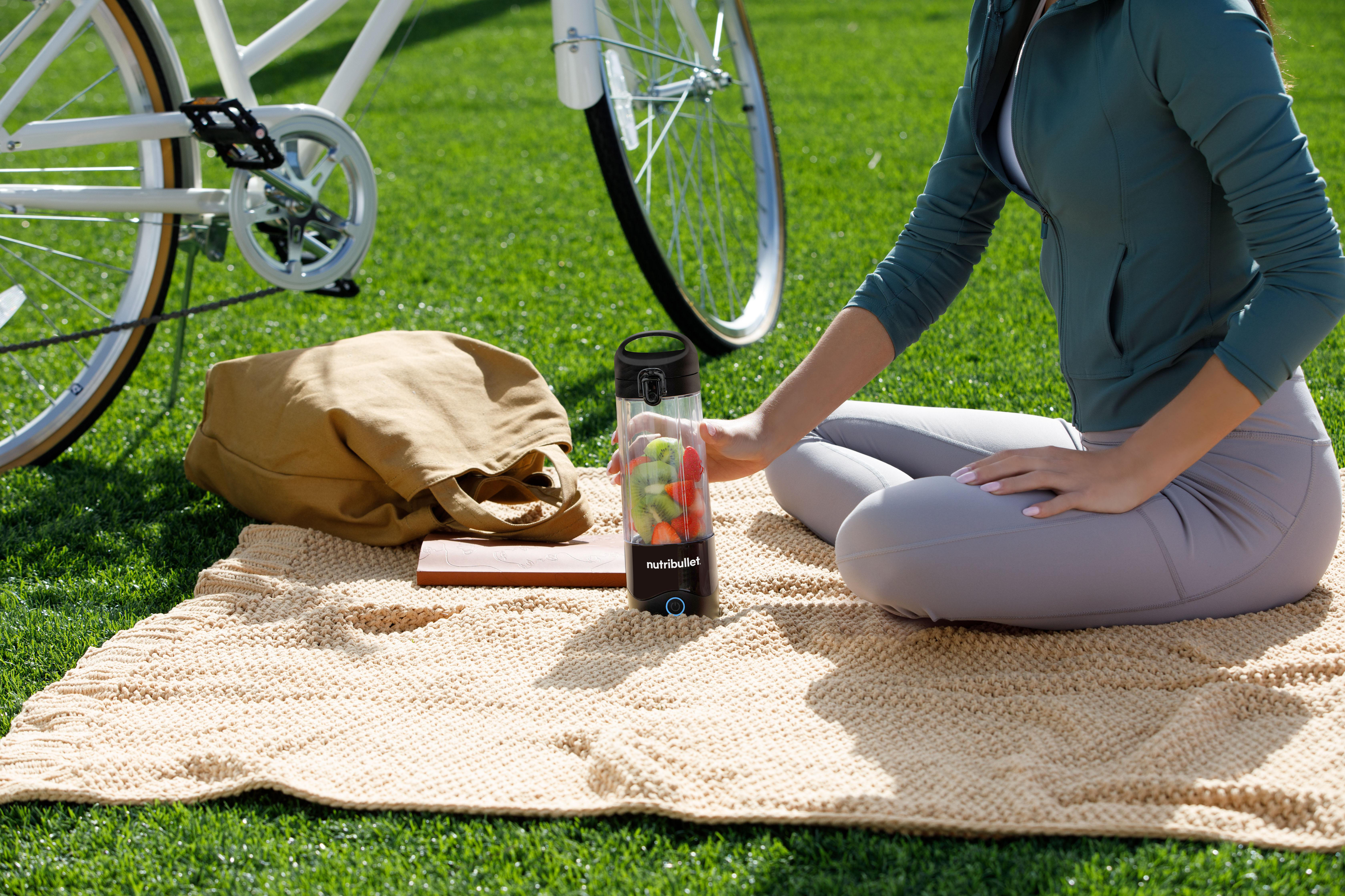 Frau auf Picknickdecke mit Mixer und Fahrrad im Hintergrund, auf grünem Grasfeld.