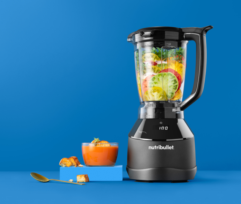 Mixer mit Zutaten, ein Glas Suppe und ein goldener Löffel auf blauem Hintergrund.