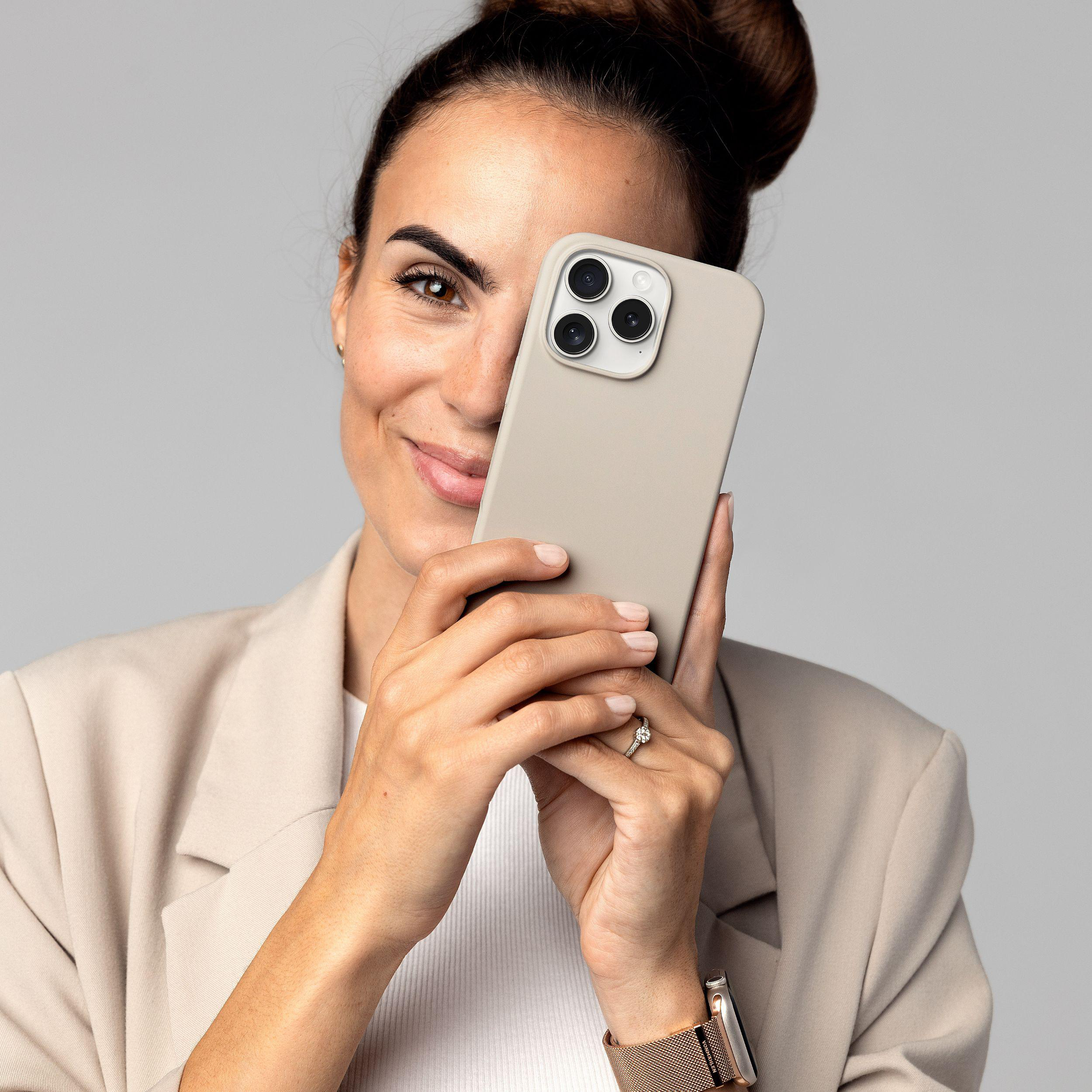Frau hält ein Telefon vor ihr Gesicht, lächelt, trägt einen Blazer. Grauer Hintergrund.