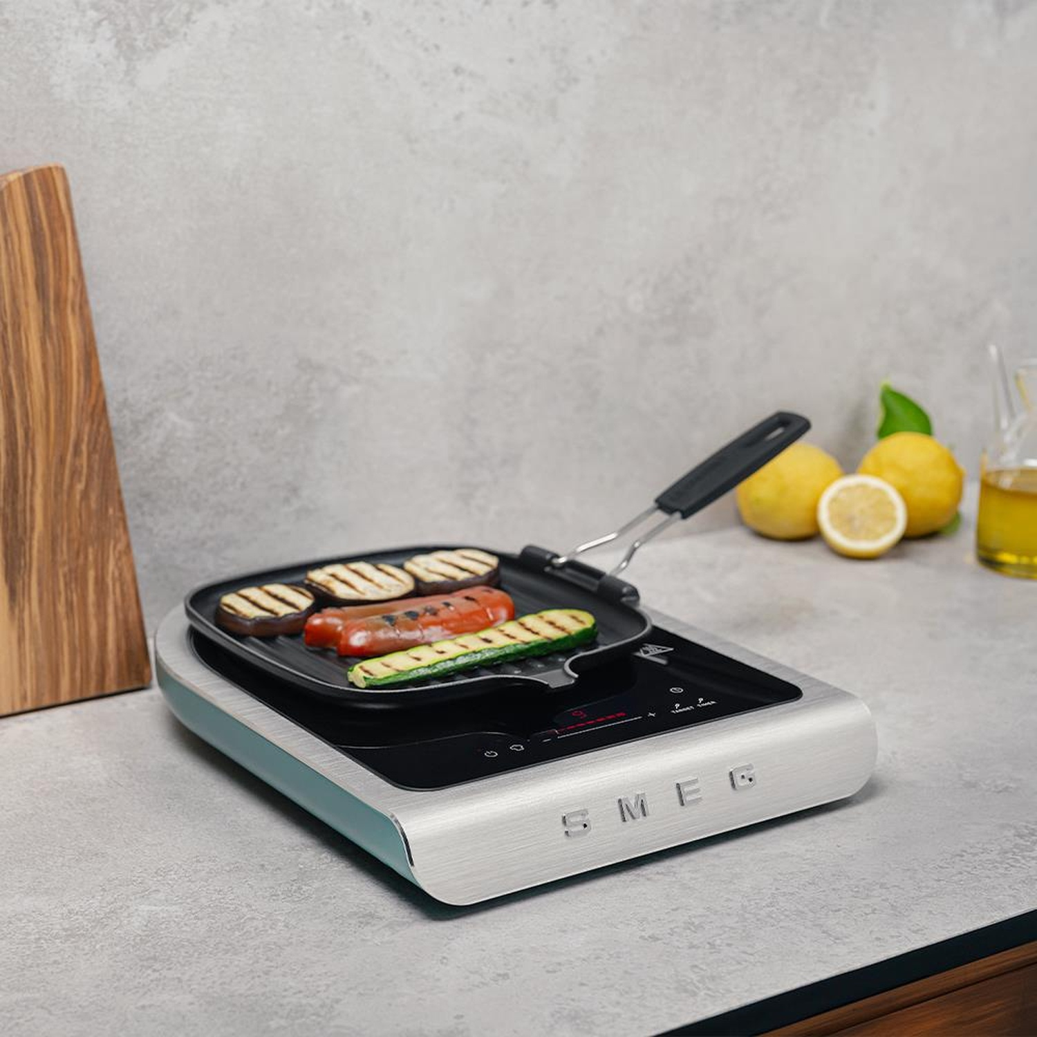 Un piano cottura a induzione SMEG con una bistecchiera sopra. La padella contiene salsicce e verdure grigliate.