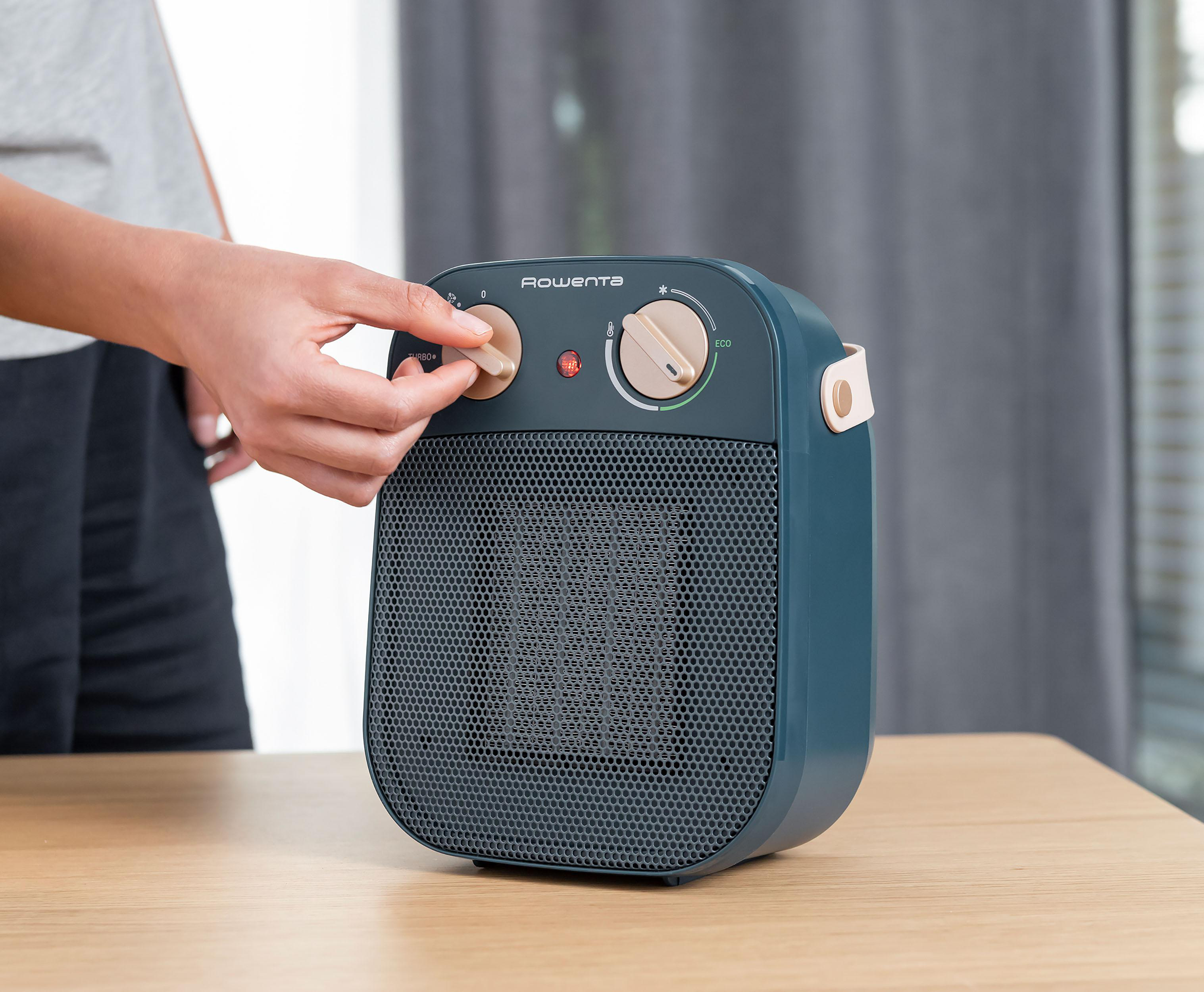 Hand stellt den Regler eines blauen Heizlüfters ein. Der Heizlüfter steht auf einem Holztisch.