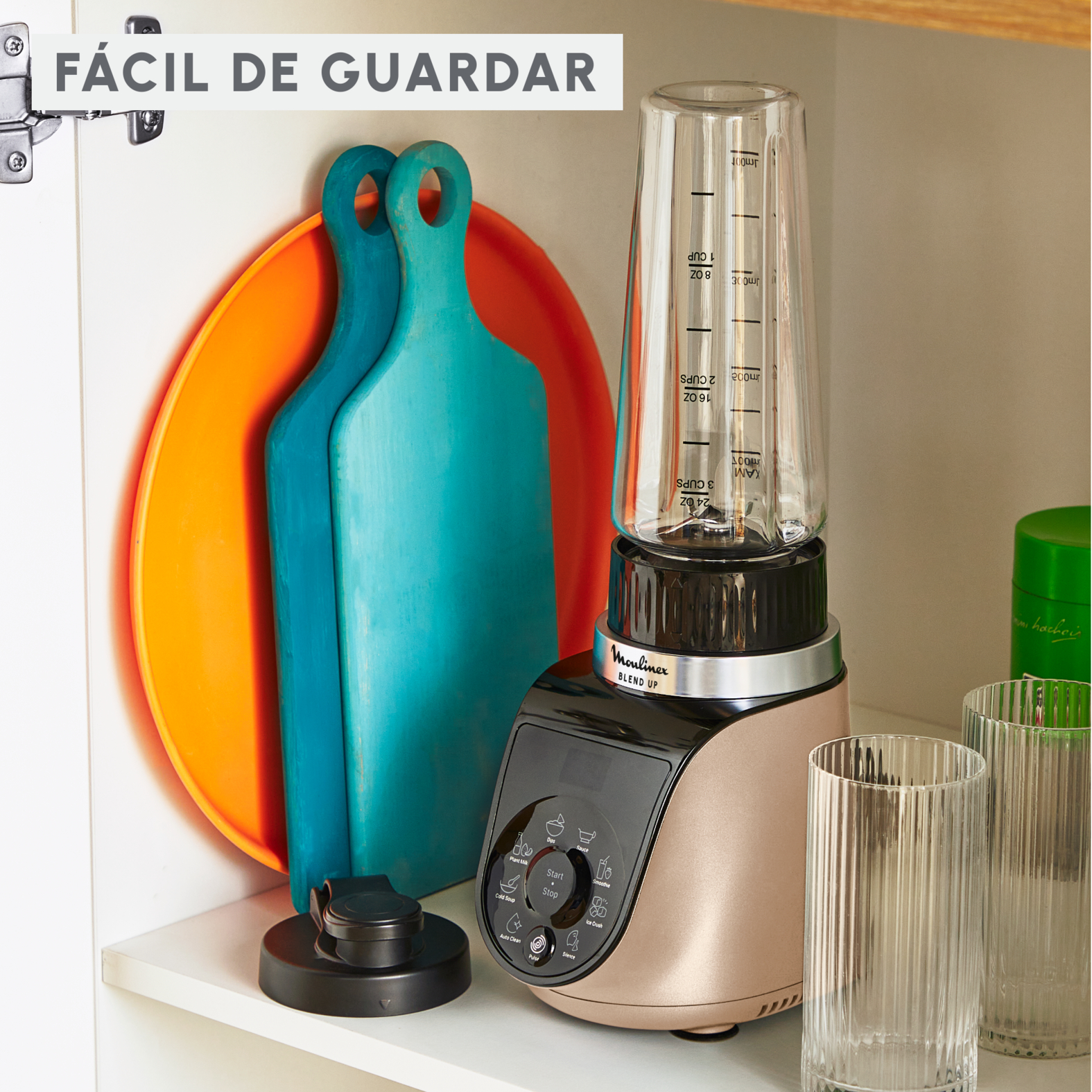Una licuadora con una taza medidora, tablas de cortar y vasos en un armario de cocina. El texto dice 'Fácil de guardar'.
