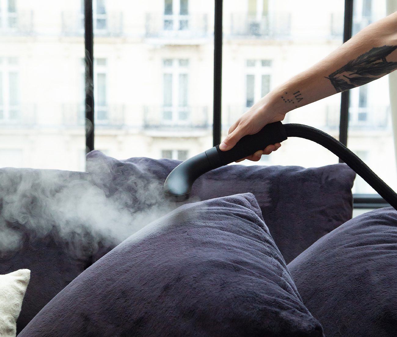 Une main vaporise un coussin de canapé violet foncé, avec une fenêtre et une ligne d'horizon en arrière-plan.
