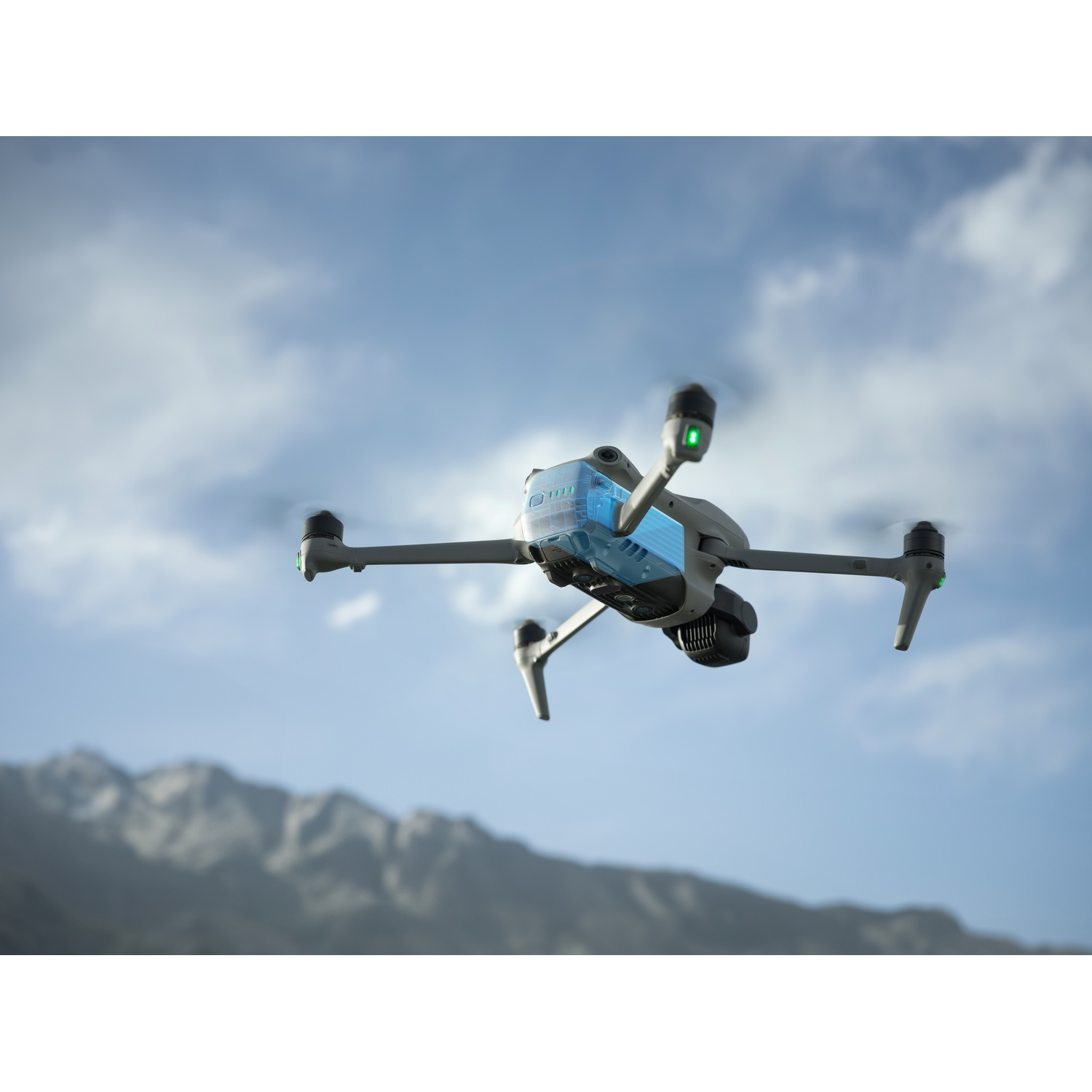 Un drone grigio in volo contro un cielo nuvoloso e montagne sullo sfondo.