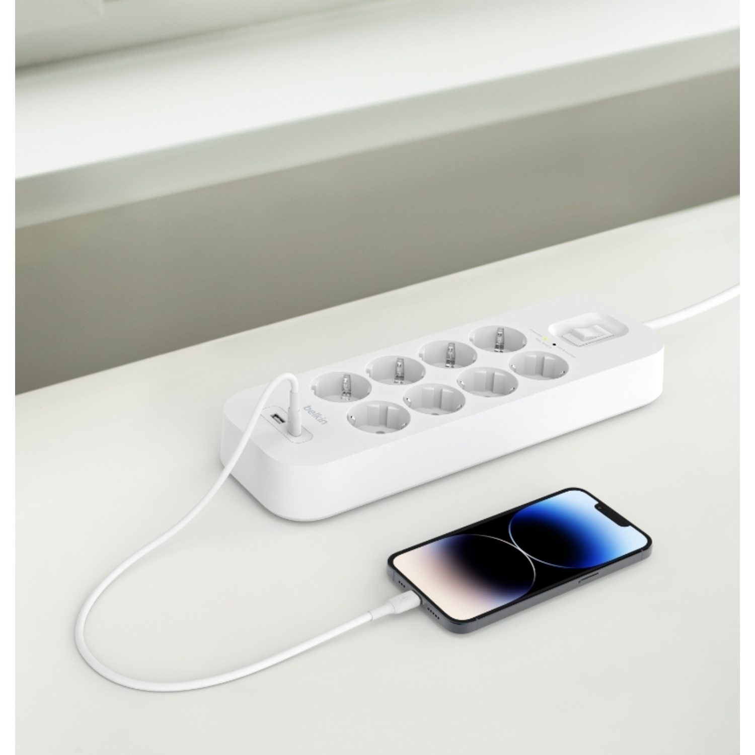 Een witte stekkerdoos met een aangesloten telefoon en een raam op de achtergrond.
