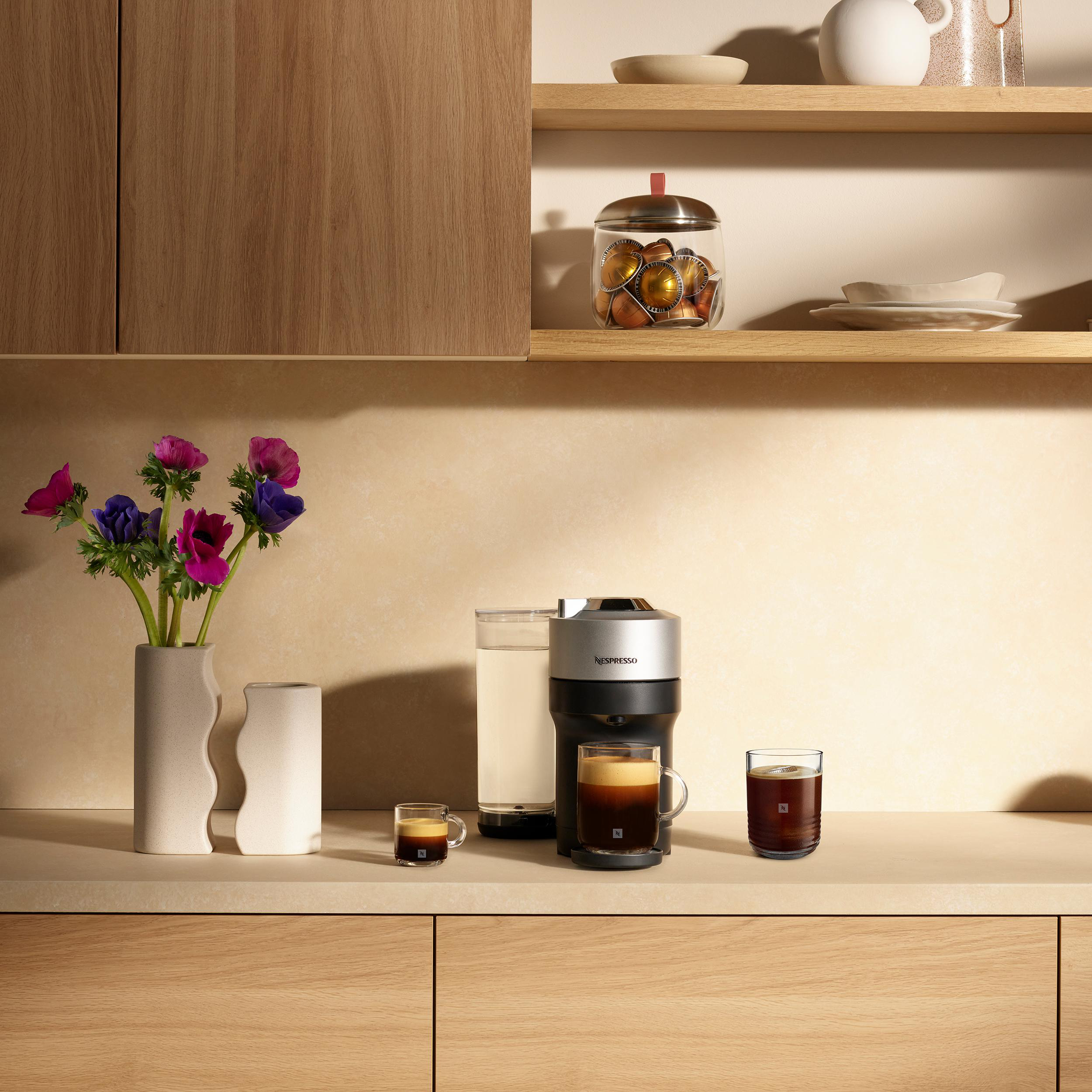 Eine Kaffeemaschine mit zwei Kaffeetassen, einer Blumenvase und Kaffeekapseln steht auf einer Theke.