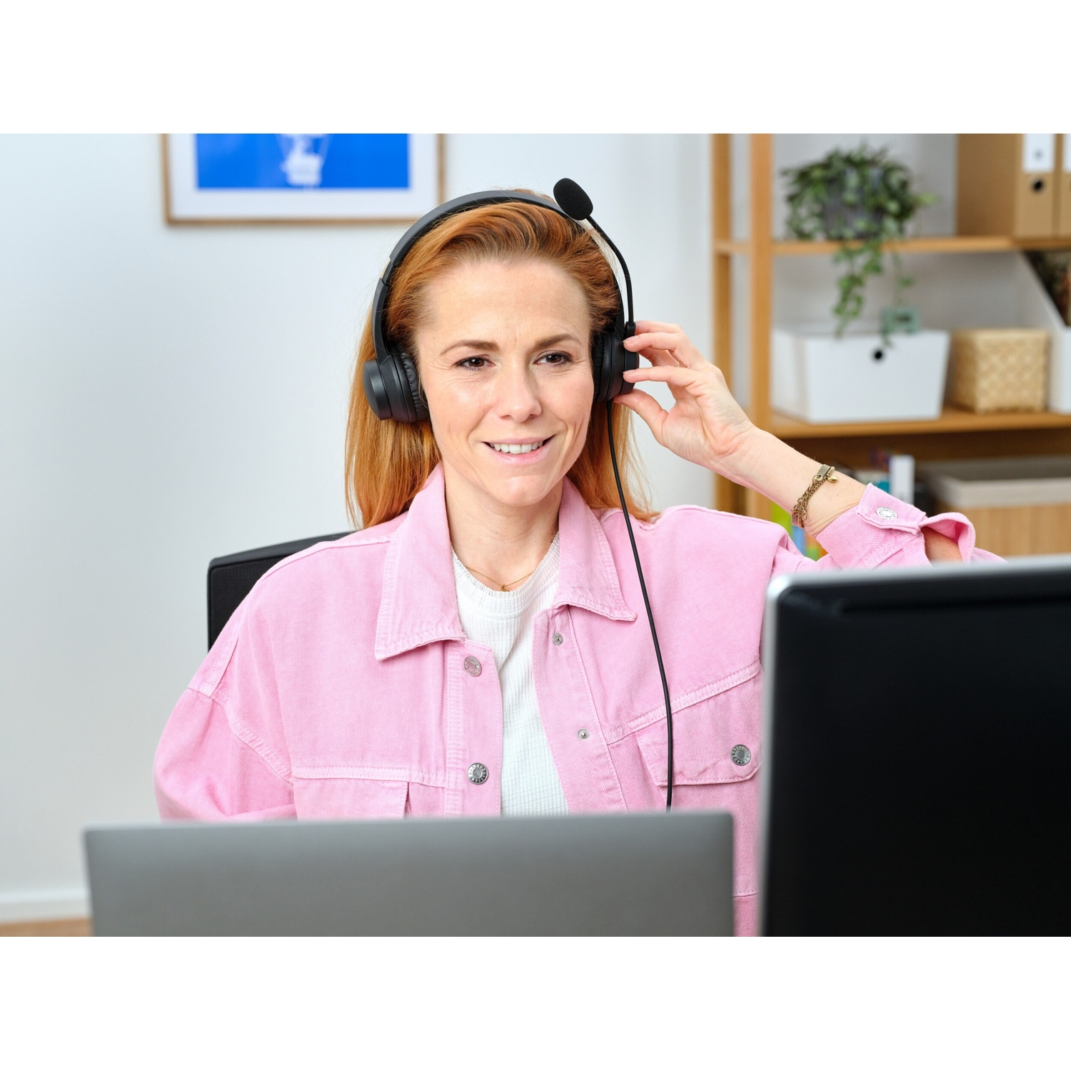 Una donna con le cuffie sorride a un laptop. Indossa una giacca rosa. Lo sfondo è un ufficio domestico.