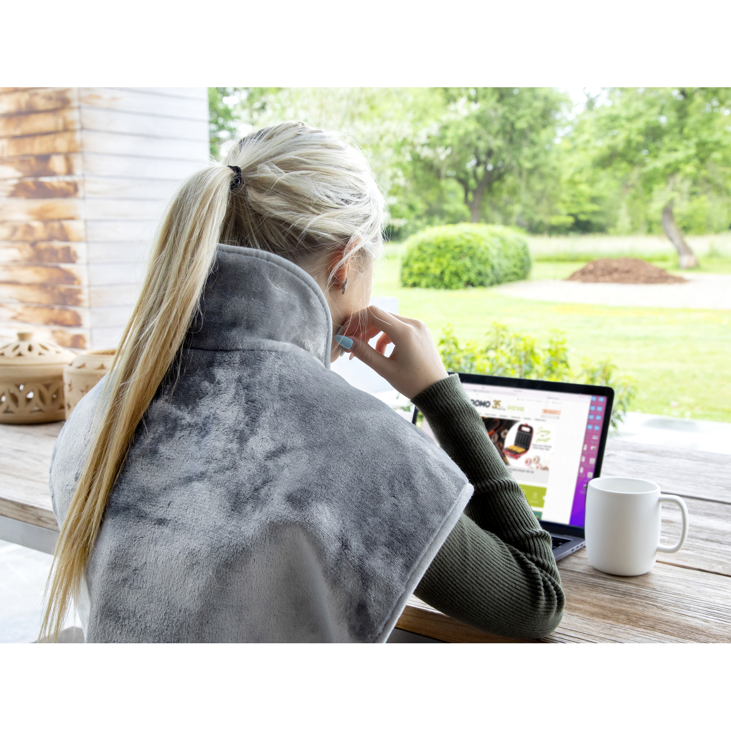 Une femme assise dehors à une table, utilisant un ordinateur portable. Un coussin chauffant autour de son cou. Tasse et jardin.