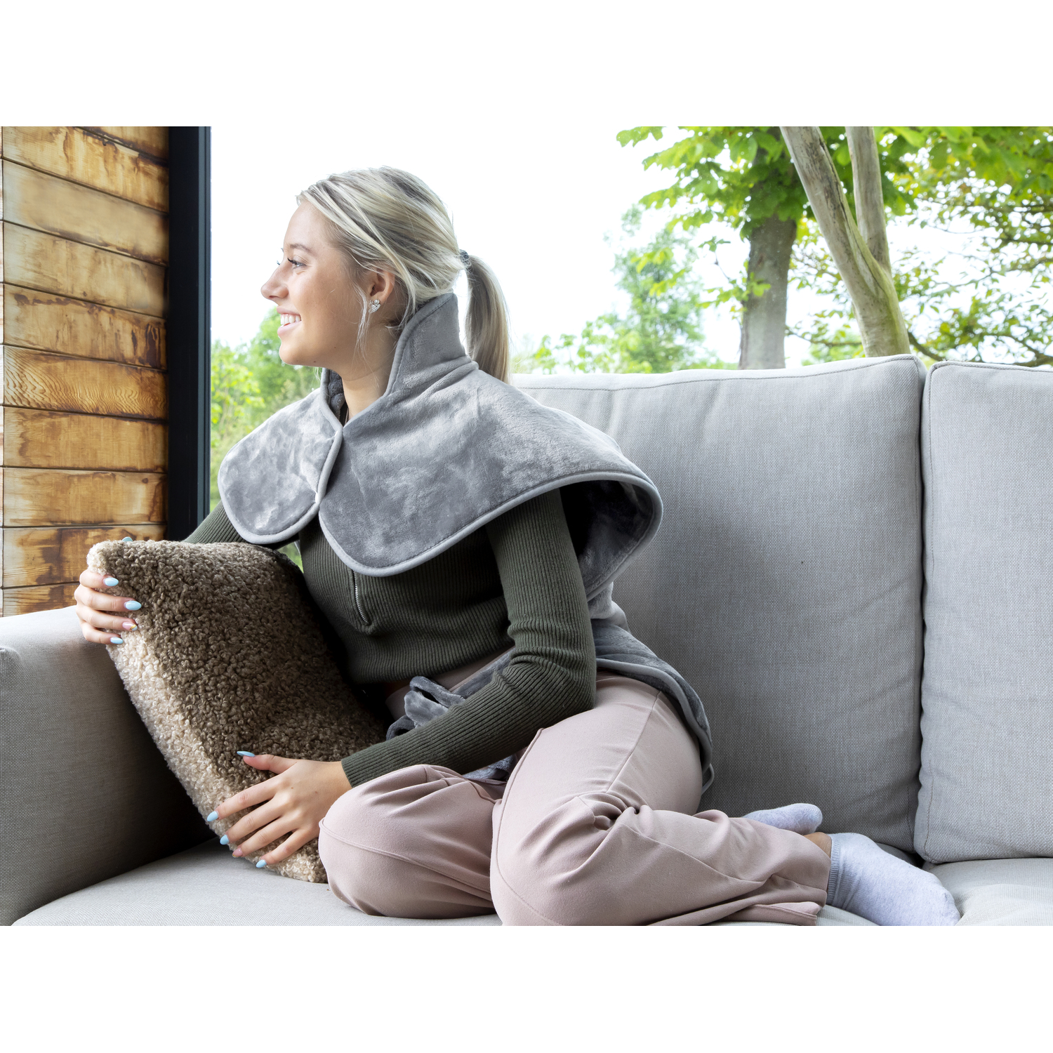 Femme portant un chauffe-nuque gris sur un canapé, regardant par la fenêtre, tenant un oreiller.