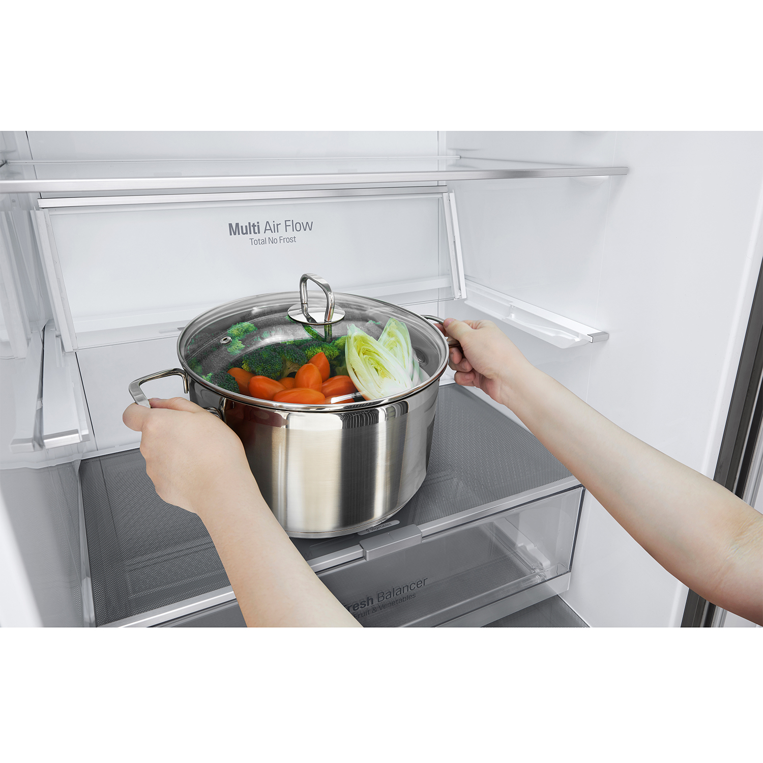 Le mani mettono una pentola di verdure in un frigorifero. La pentola contiene carote, broccoli e lattuga. Il frigo è bianco.