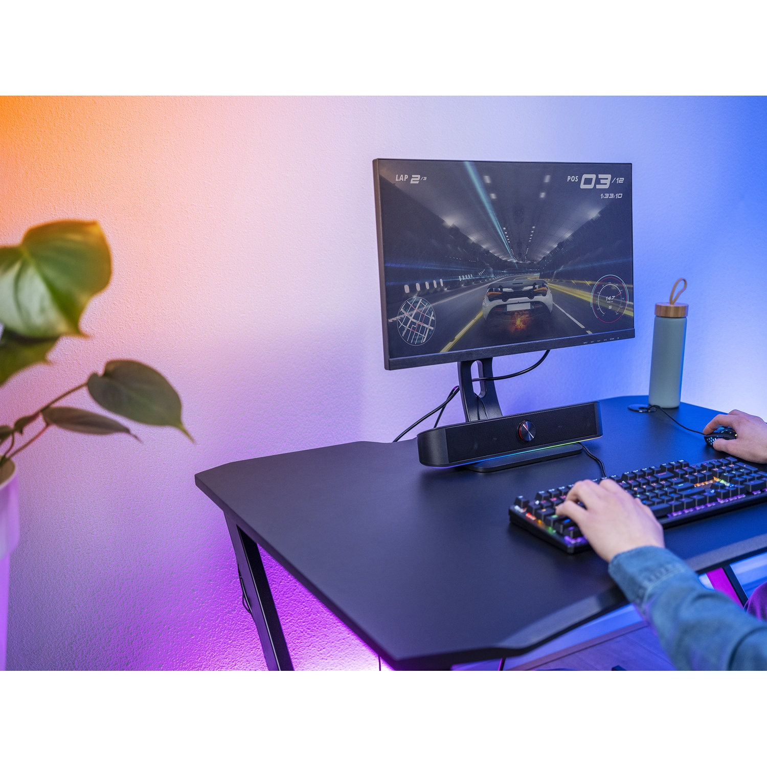 Persona che gioca su un computer con un gioco di corse visualizzato sul monitor. Sono visibili una scrivania, una tastiera, un altoparlante e una bottiglia d'acqua.