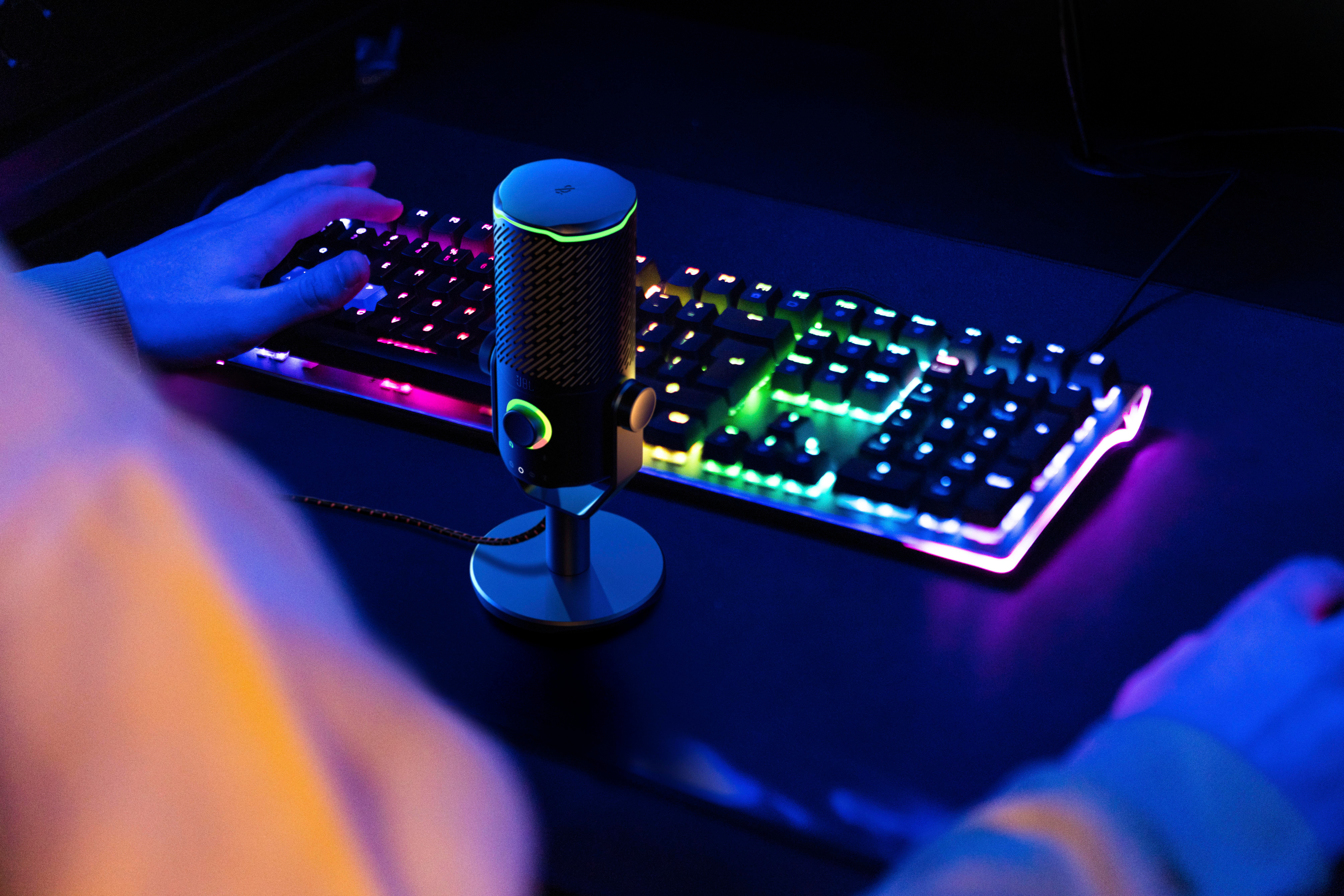 Gaming-Setup mit Mikrofon, Tastatur mit beleuchteten Tasten und tippenden Händen. Dunkle Umgebung.
