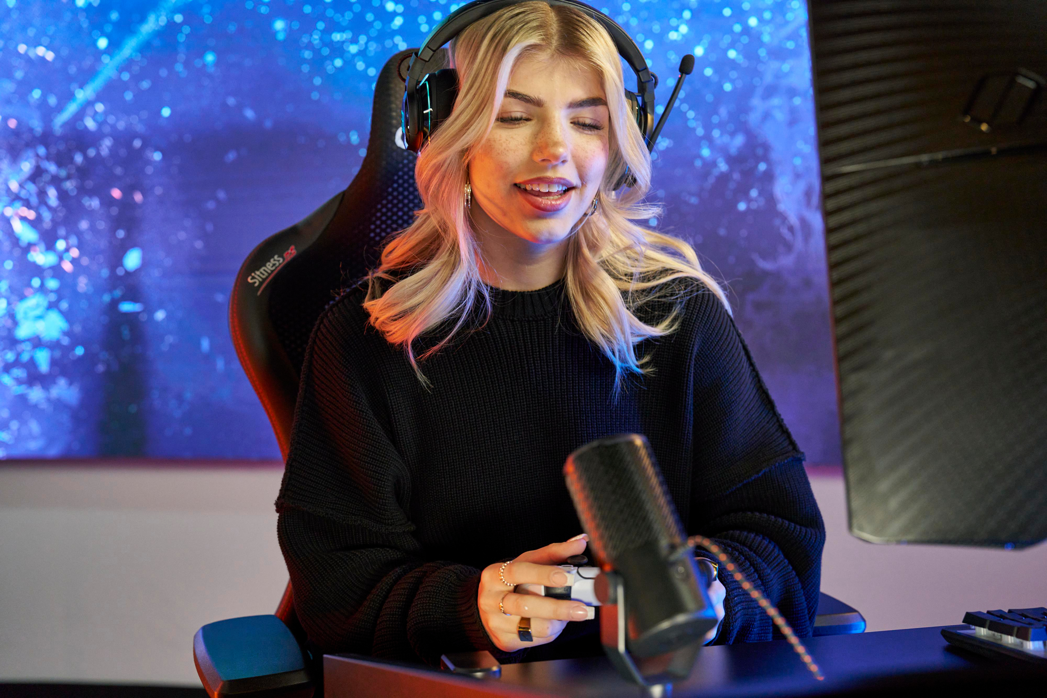 Blonde Frau mit Kopfhörern und Mikrofon spielt, lächelt, mit Controller in der Hand.