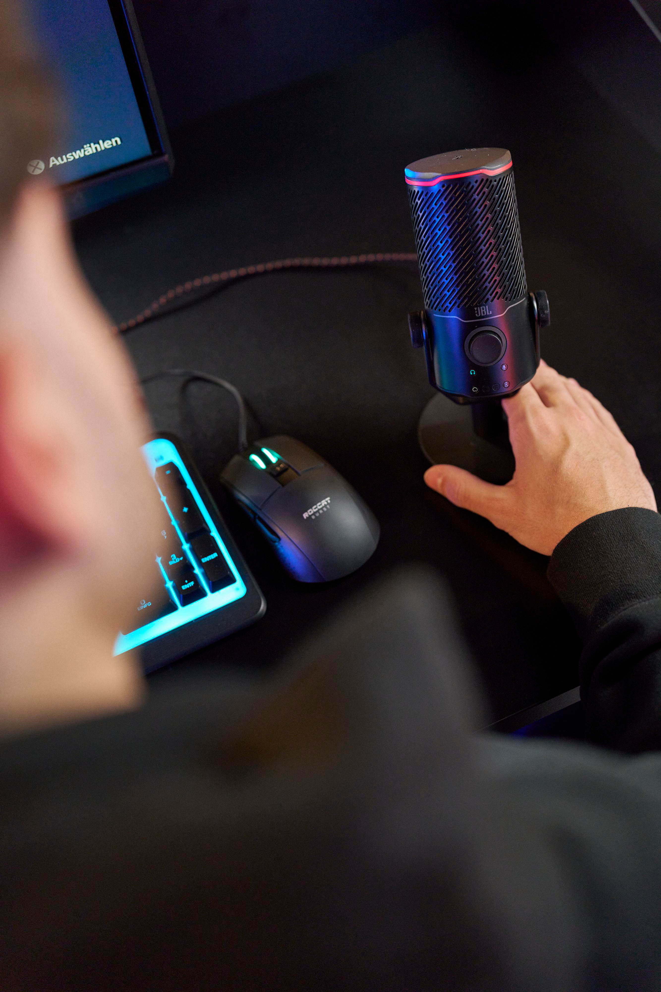 Die Hand einer Person, die ein schwarzes JBL-Mikrofon auf einem Schreibtisch mit Maus und Tastatur einstellt.